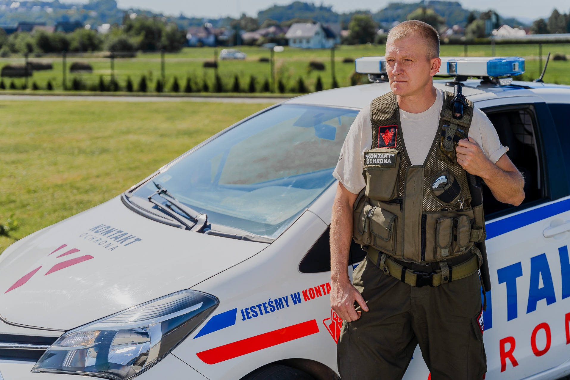 Mężczyzna w kamizelce ochronnej z krótkofalówką, stojący obok oznakowanego samochodu firmy ochroniarskiej z niebieskimi światłami ostrzegawczymi na dachu i napisem 'KONTAKT OCHRONA' na masce.