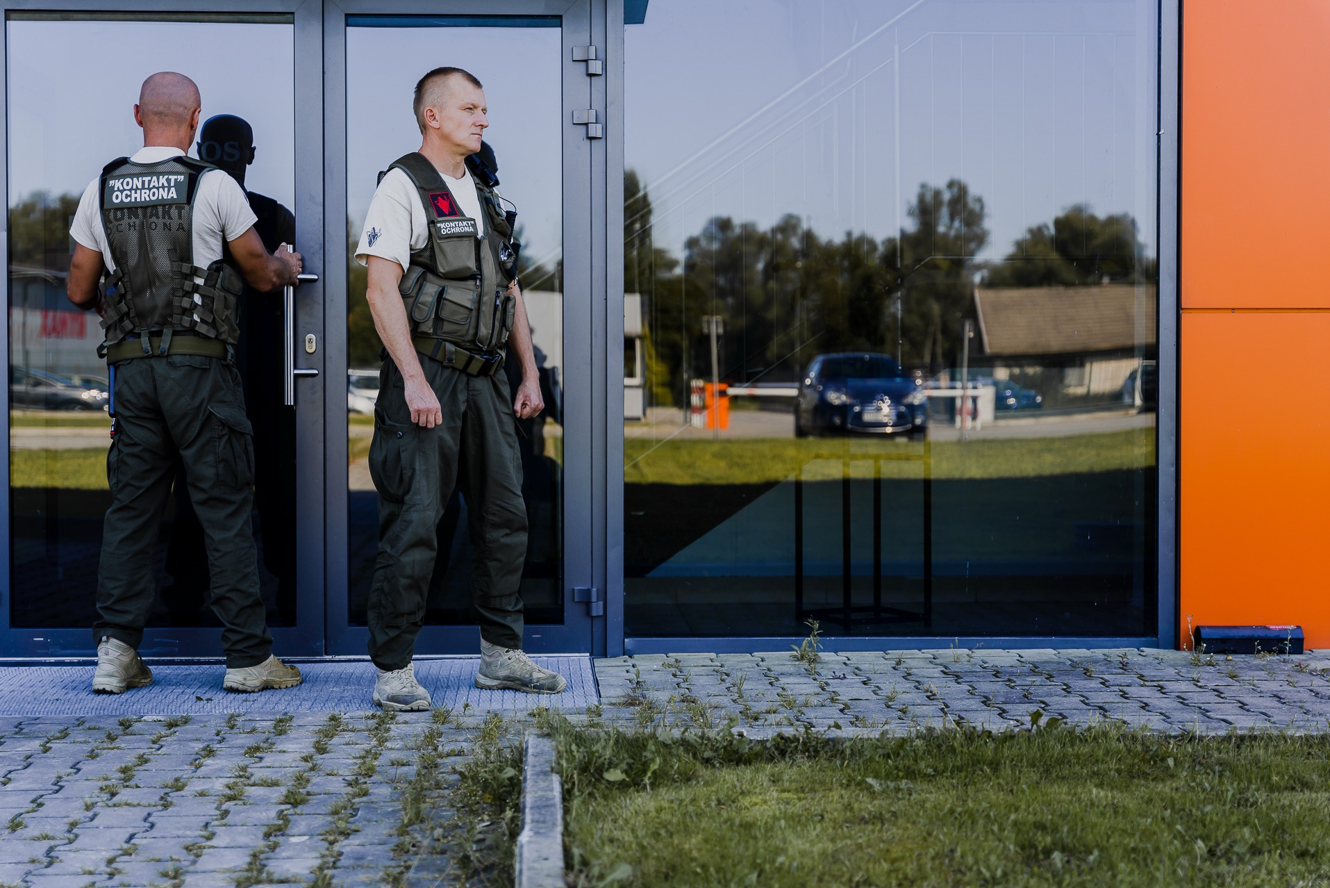 Dwóch ochroniarzy w mundurach i kamizelkach taktycznych stoi przed nowoczesnym budynkiem z przeszklonymi drzwiami, w których odbija się zaparkowany samochód i otoczenie.