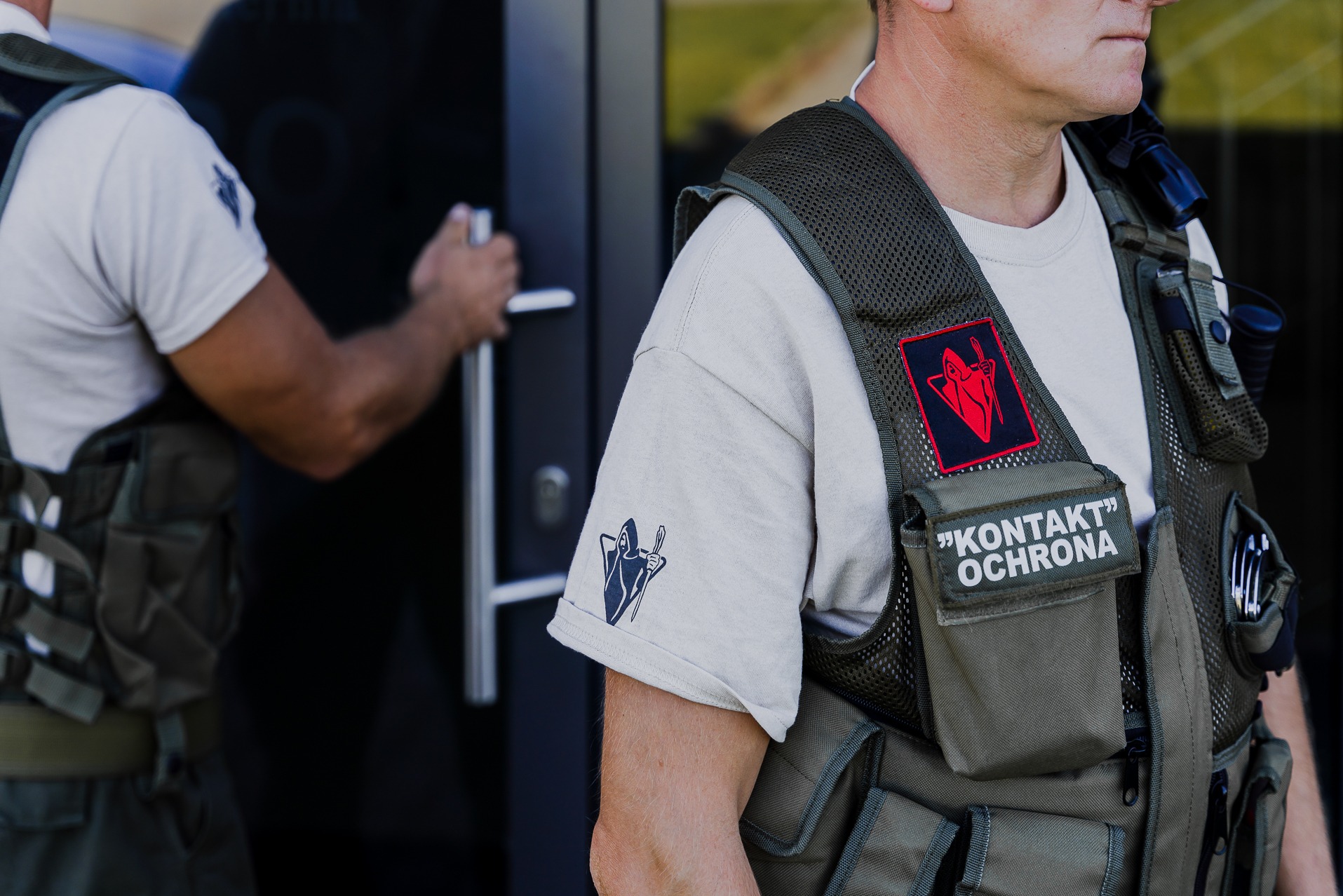 Fragment umundurowania pracownika ochrony z widocznym logotypem firmy na ramieniu i kamizelce taktycznej, otwierającego drzwi wejściowe.