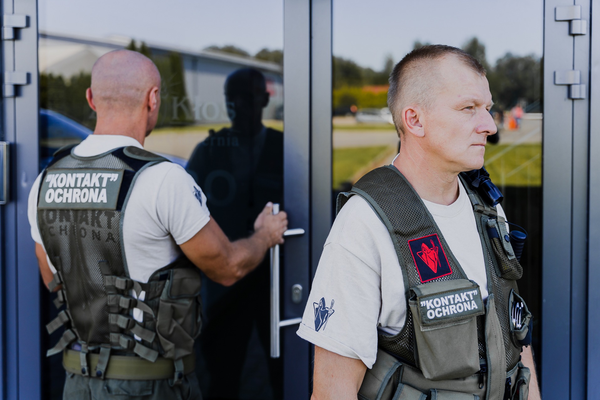 Dwóch ochroniarzy w kamizelkach z napisem 'KONTAKT OCHRONA' wchodzi do budynku o szklanych drzwiach, jeden z nich otwiera drzwi, drugi patrzy w prawo.