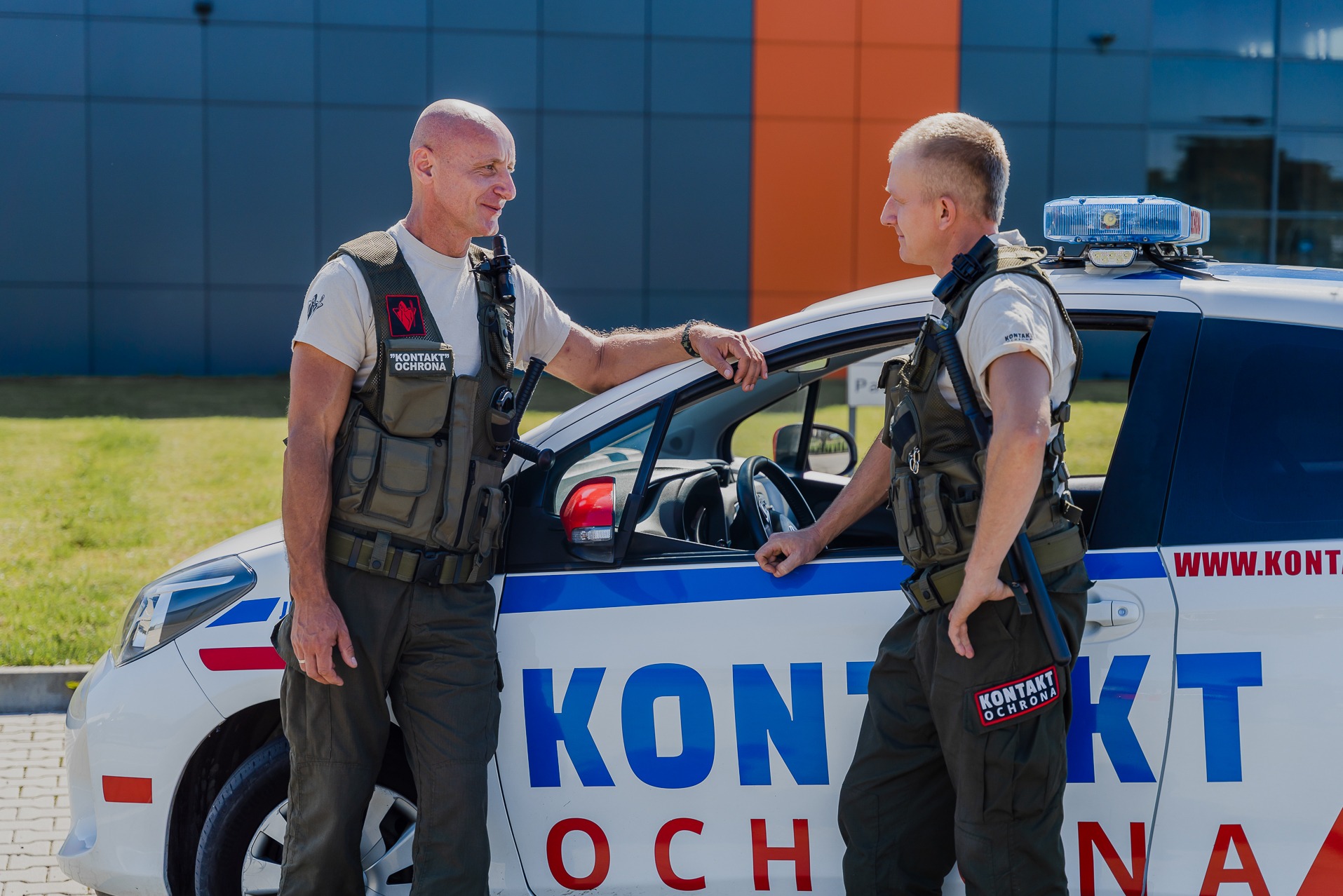 Dwóch umundurowanych ochroniarzy w kamizelkach taktycznych rozmawia opierając się o oznakowany samochód patrolowy z niebieskim kogutem na dachu, na tle budynku z niebiesko-pomarańczową elewacją.