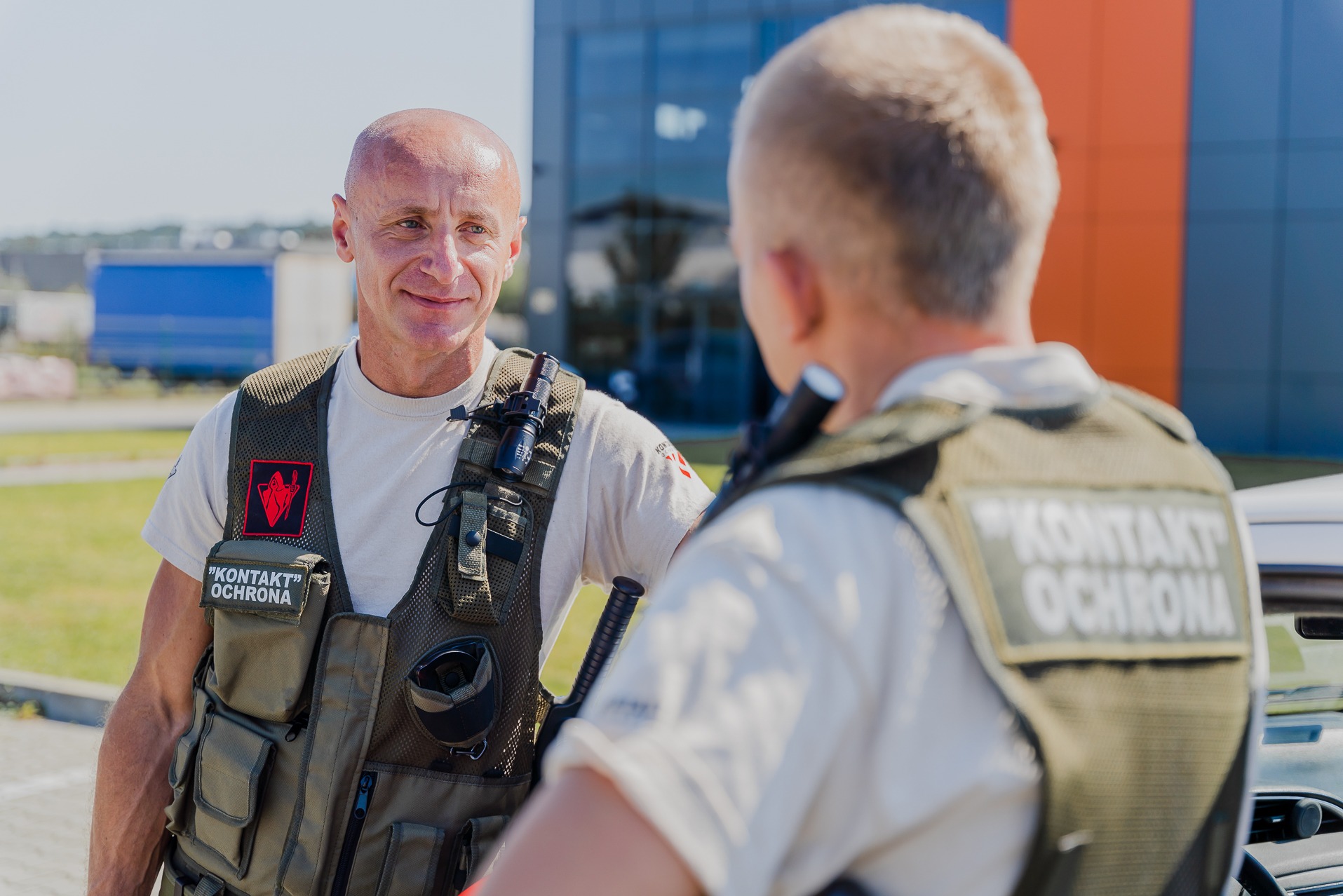 Dwóch ochroniarzy firmy 'Kontakt Ochrona' w kamizelkach taktycznych rozmawia na tle nowoczesnego budynku i zaparkowanego samochodu.