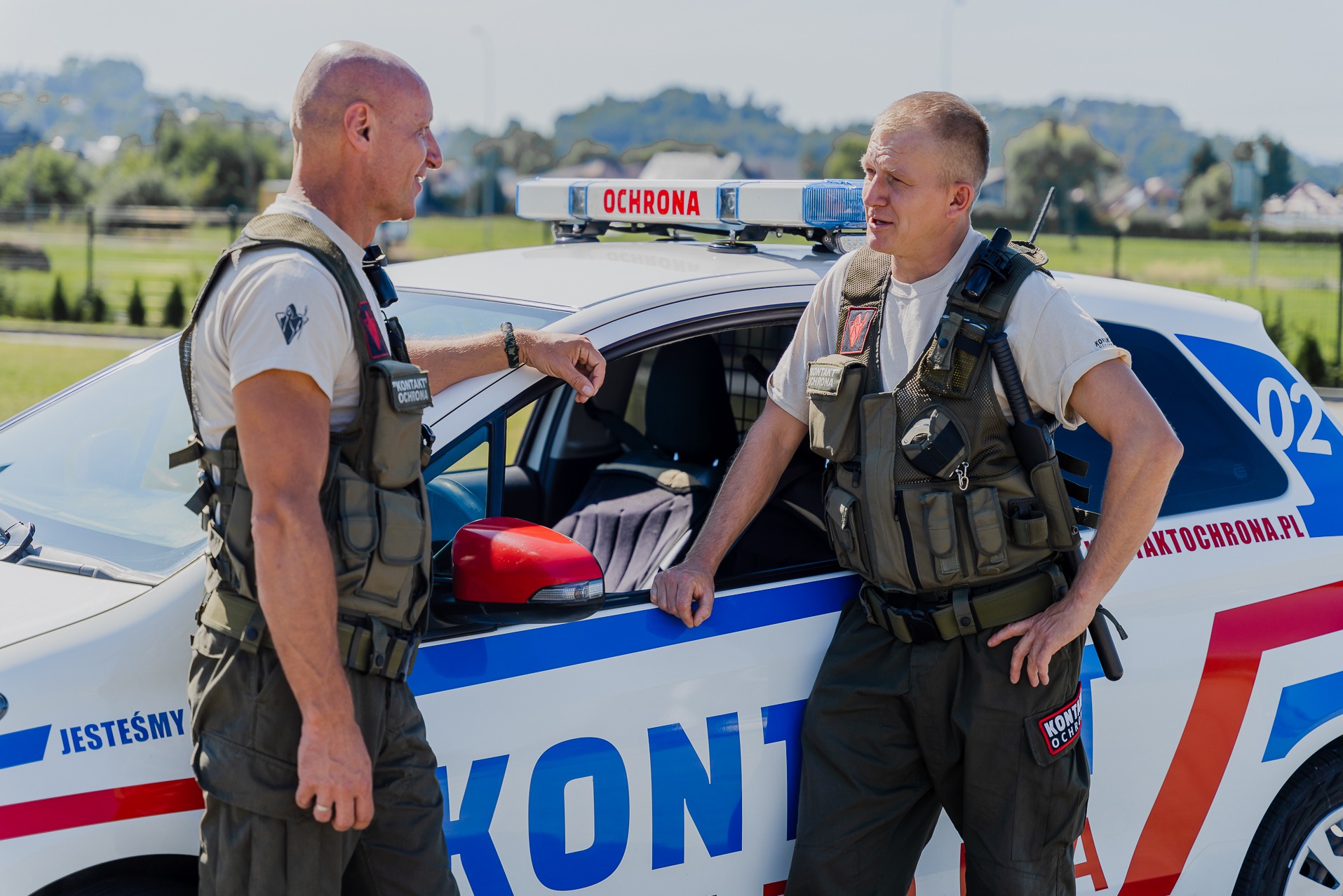 Dwóch umundurowanych ochroniarzy rozmawia obok oznakowanego radiowozu z napisem 'OCHRONA' i niebiesko-czerwonymi pasami, widoczne logo firmy na mundurach i samochodzie.
