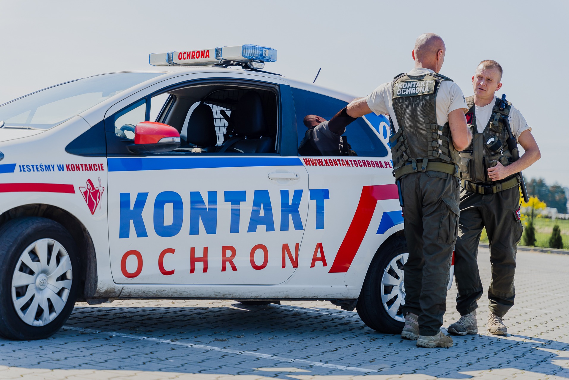 Dwóch ochroniarzy w kamizelkach stoi obok oznakowanego samochodu patrolowego z napisem KONTAKT OCHRONA, jeden z nich opiera się o otwarte okno pojazdu.