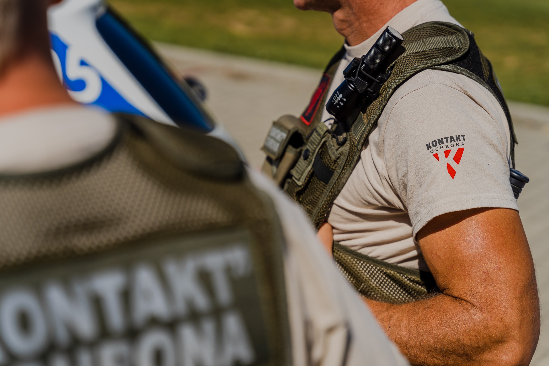 Dwóch ochroniarzy w kamizelkach taktycznych i koszulkach z logo firmy ochroniarskiej, jeden z latarką na ramieniu, stojący obok pojazdu.