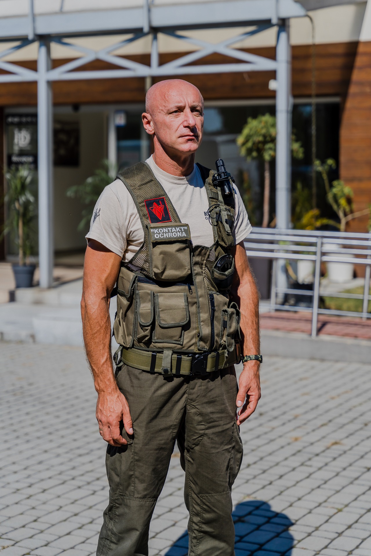 Mężczyzna w uniformie ochroniarza z kamizelką taktyczną i radiotelefonem stoi na zewnątrz budynku z przeszkloną fasadą.