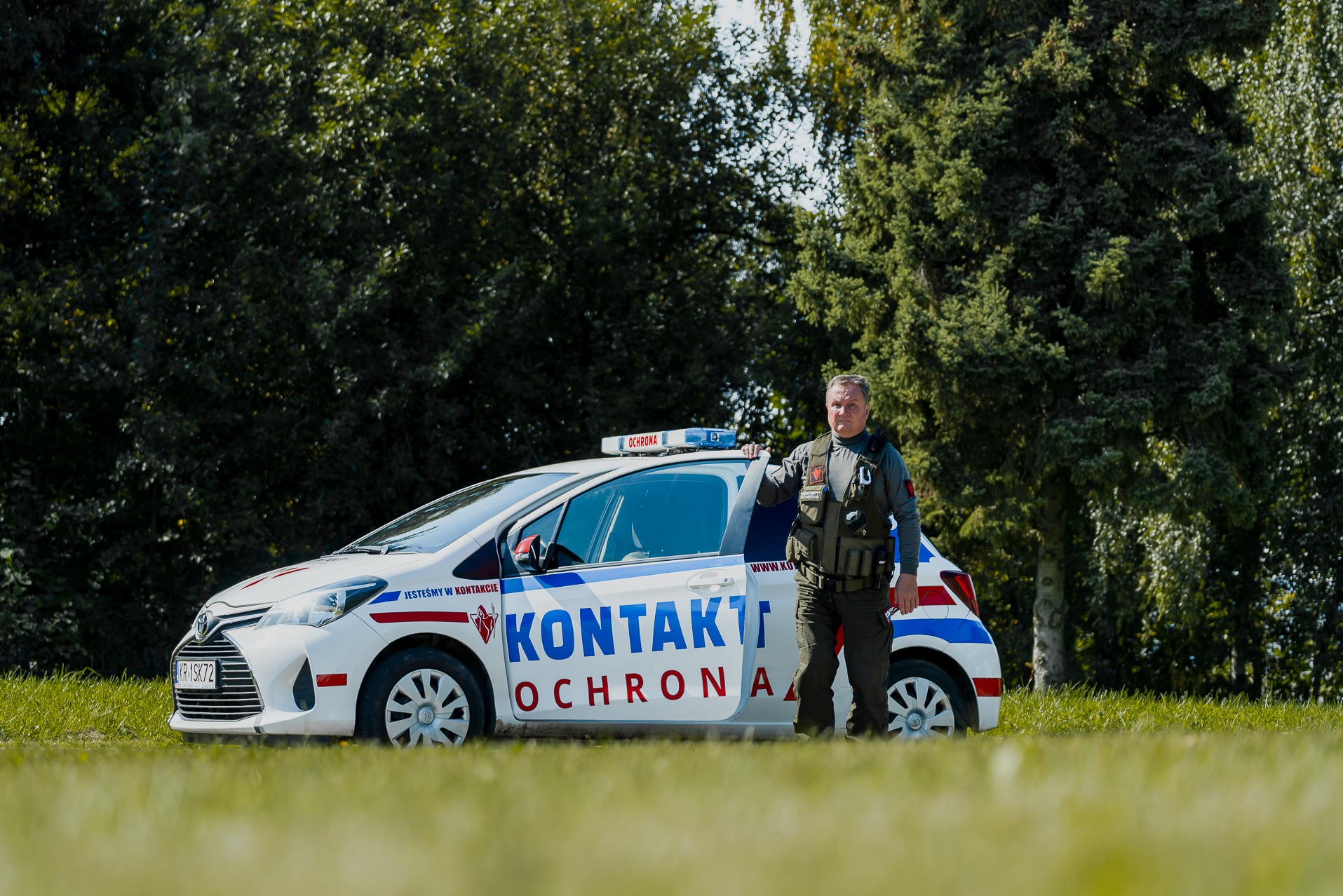 Pracownik ochrony w kamizelce taktycznej stoi obok oznakowanego samochodu patrolowego firmy ochroniarskiej na tle zieleni.