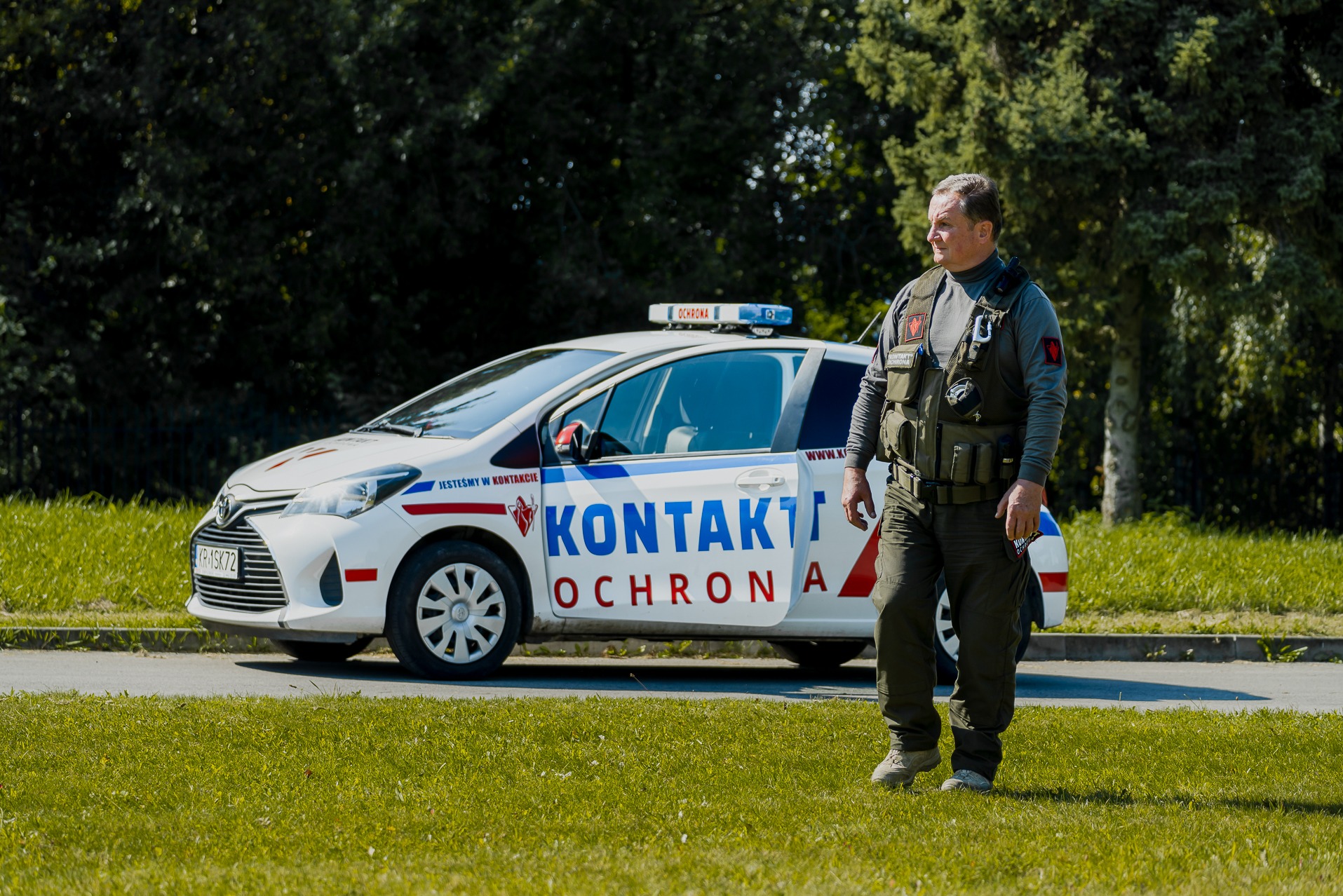 Mężczyzna w kamizelce taktycznej stoi na trawniku obok oznakowanego samochodu osobowego z napisem KONTAKT OCHRONA i niebieskim kogutem na dachu.