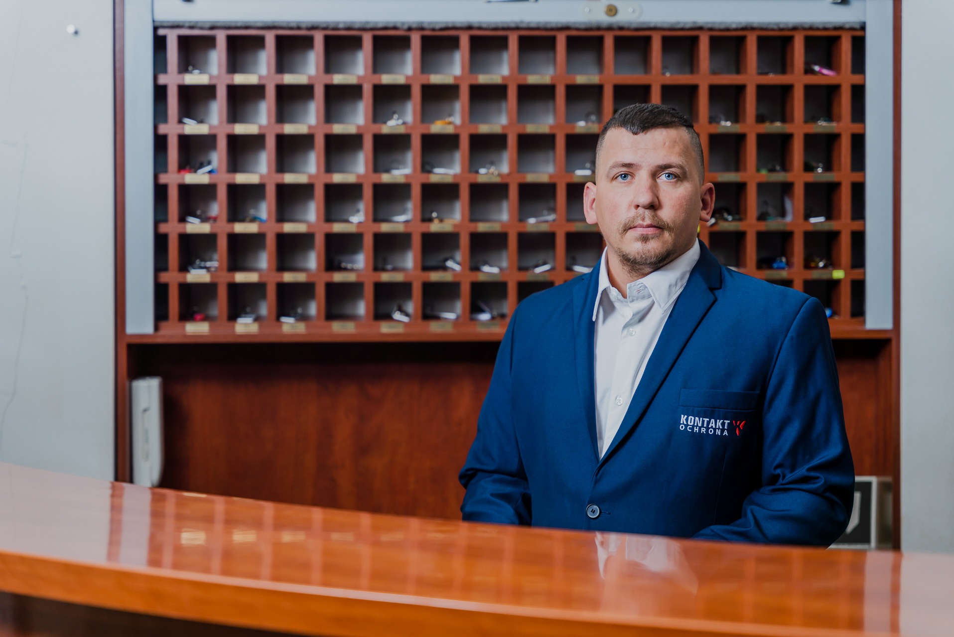 Mężczyzna w niebieskiej marynarce z logo firmy ochroniarskiej 'KONTAKT Ochrona' stojący za recepcją z drewnianymi przegródkami na klucze w tle.
