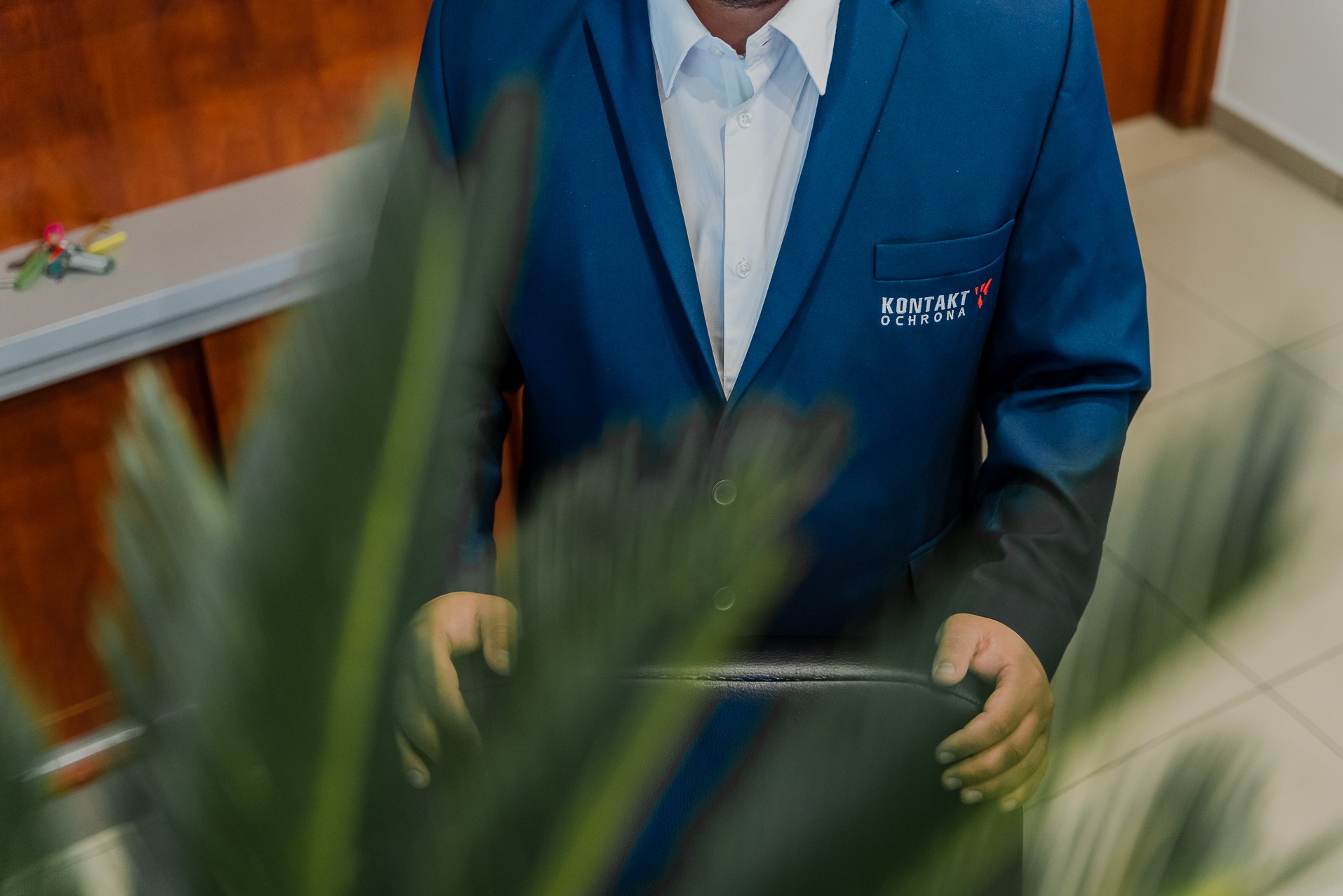 Mężczyzna w niebieskim garniturze z logo firmy ochroniarskiej 'Kontakt Ochrona', siedzący za biurkiem, częściowo zasłonięty rośliną doniczkową.