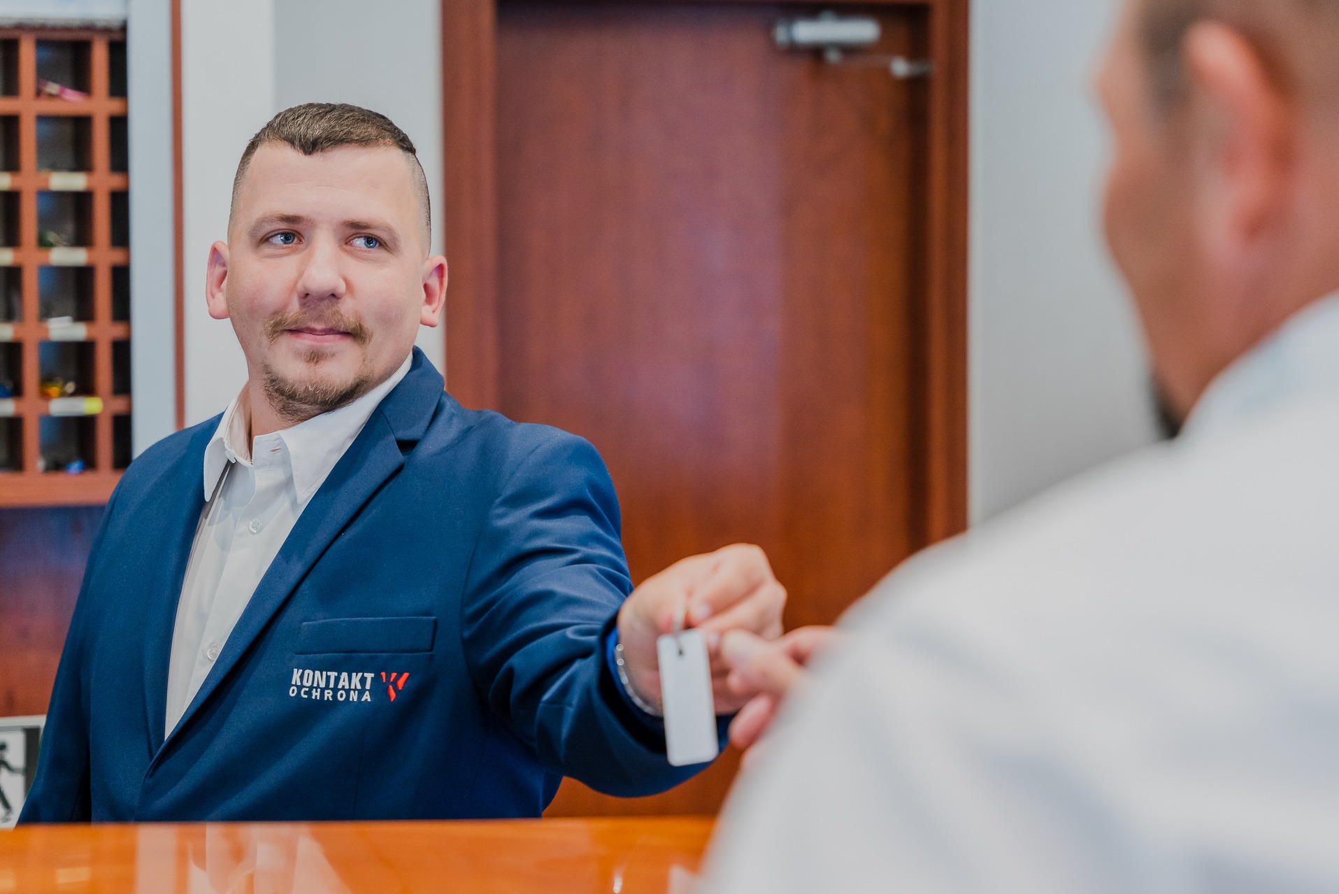 Pracownik ochrony w granatowym garniturze z logo firmy 'Kontakt Ochrona' wręcza kartę dostępu osobie poza kadrem, na tle recepcji.