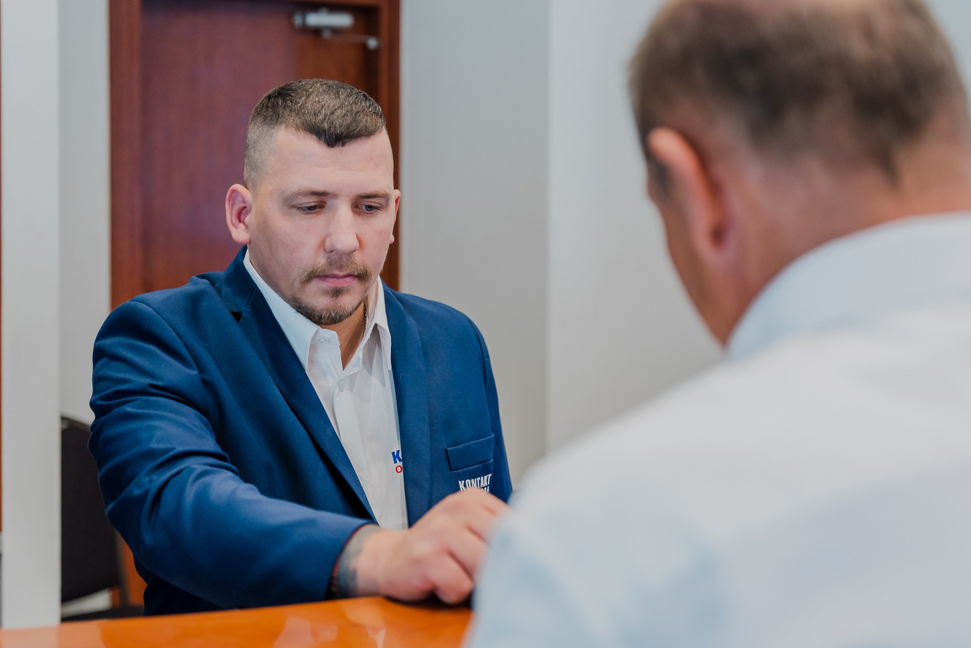 Mężczyzna w niebieskiej marynarce z logo firmy ochroniarskiej na kieszeni, rozmawia z klientem przy pomarańczowym biurku.