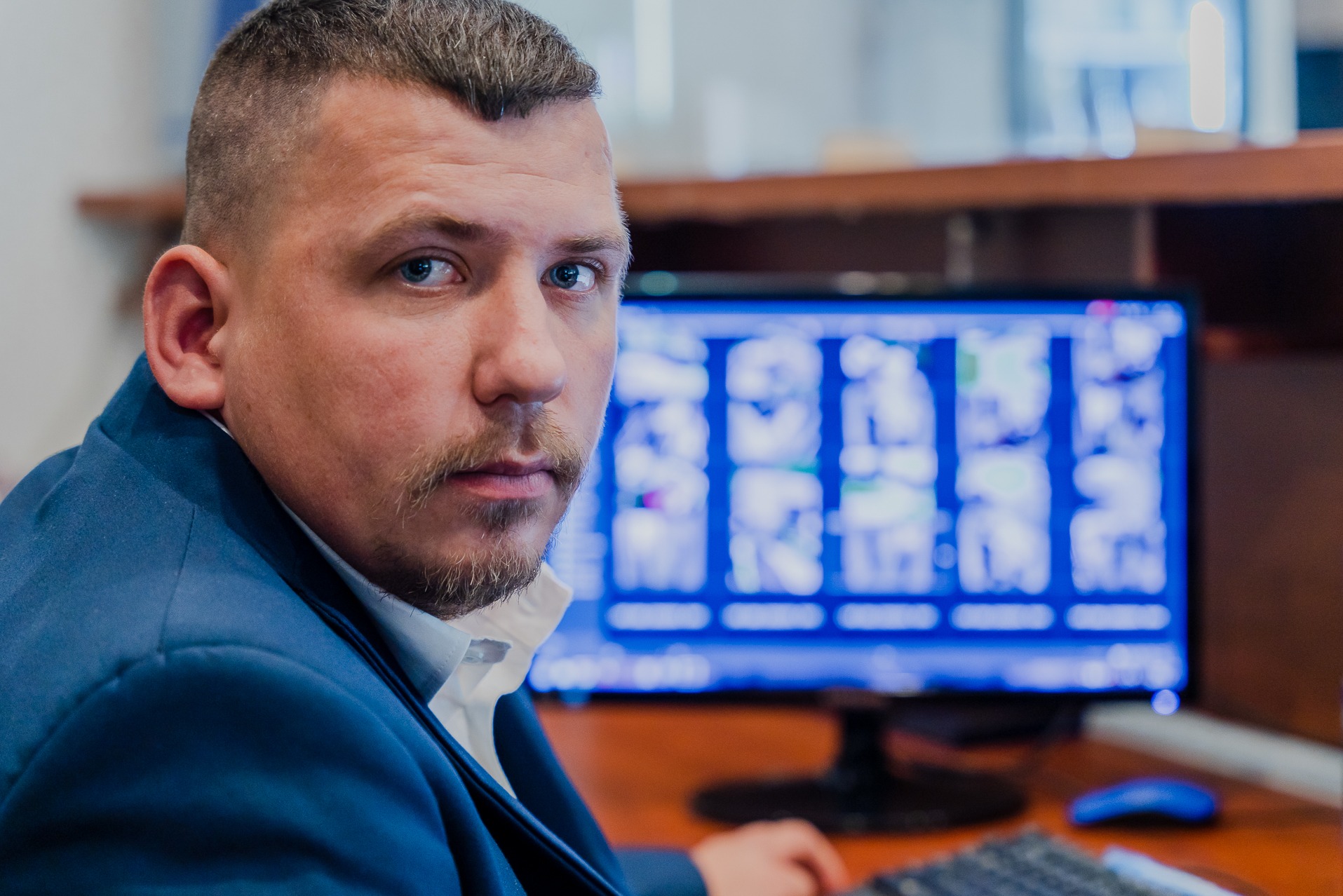 Mężczyzna w niebieskiej marynarce, patrzący w kamerę, z ekranem monitoringu w tle, prezentującym obraz z wielu kamer.