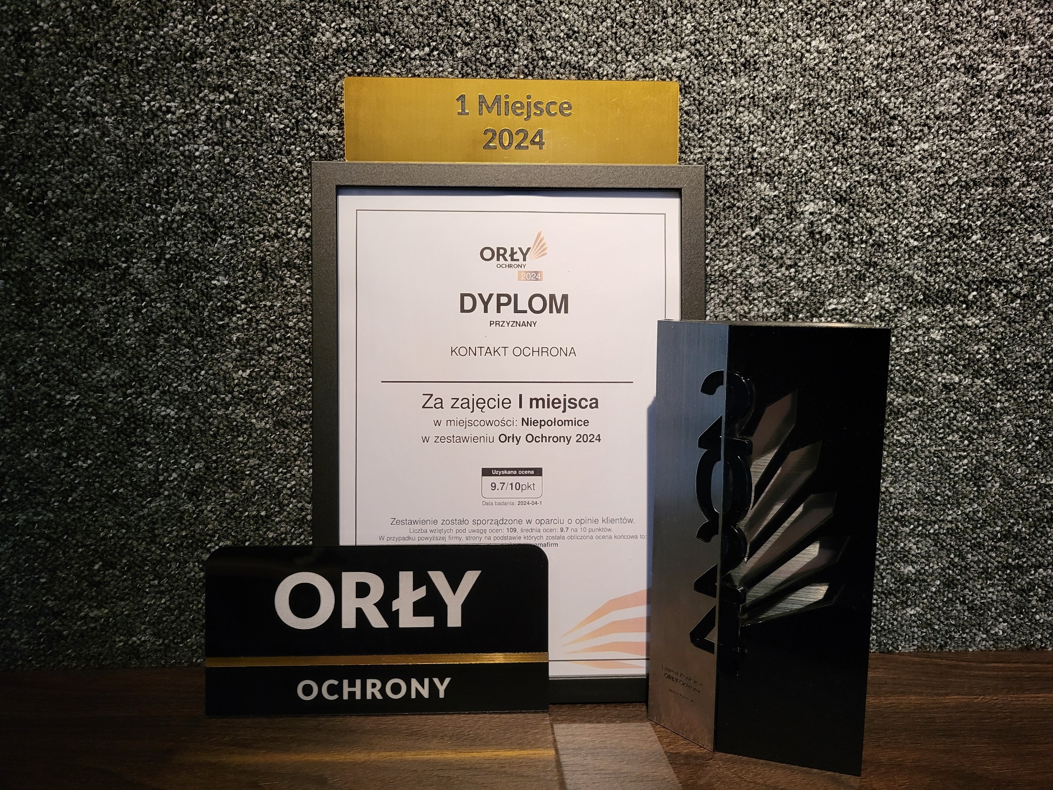 Dyplom 'Orły Ochrony 2024' za zajęcie 1 miejsca w Niepołomicach, z oceną 9.7/10, obok statuetka i tabliczka z logo firmy ochroniarskiej.