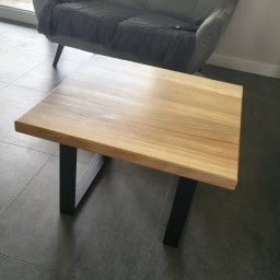 Glamour Wood - Nowoczesny stolik kawowy z jasnego drewna z czarnymi, geometrycznymi nogami, stojący na ciemnych płytkach przed szarą sofą i oknem z widokiem na zieleń.