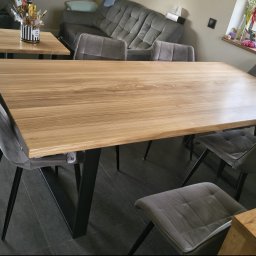 Glamour Wood - Duży stół jadalniany z blatem z jasnego drewna o naturalnej, nieregularnej krawędzi i czarnymi, trapezowymi nogami, w otoczeniu szarych krzeseł w nowoczesnym wnętrzu.