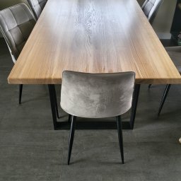 Glamour Wood - Duży stół jadalniany z jasnego drewna o wyraźnym usłojeniu, wsparty na ciemnych, geometrycznych nogach. Wokół szare, welurowe krzesła. W tle okno z zielenią i kolorowymi figurkami na parapecie.