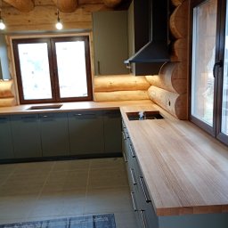 Glamour Wood - Nowoczesna kuchnia z ciemnozielonymi szafkami i jasnymi drewnianymi blatami, harmonijnie wkomponowana w wnętrze z bali. Widoczny zlew, okna i oświetlenie podszafkowe.