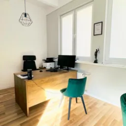 Jasne biuro: drewniane biurko z komputerem, drukarką i telefonem, czarne krzesło biurowe, zielone krzesło dla klienta, statuetka Temidy na parapecie, dyplom w ramce na ścianie, nowoczesna lampa...