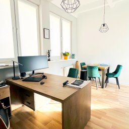 Kancelaria Adwokacka Adwokat Tomasz Siwek - Jasne biuro z drewnianym biurkiem, komputerem, dokumentami i kalkulatorem, w tle stół z zielonymi krzesłami i geometryczne lampy wiszące.