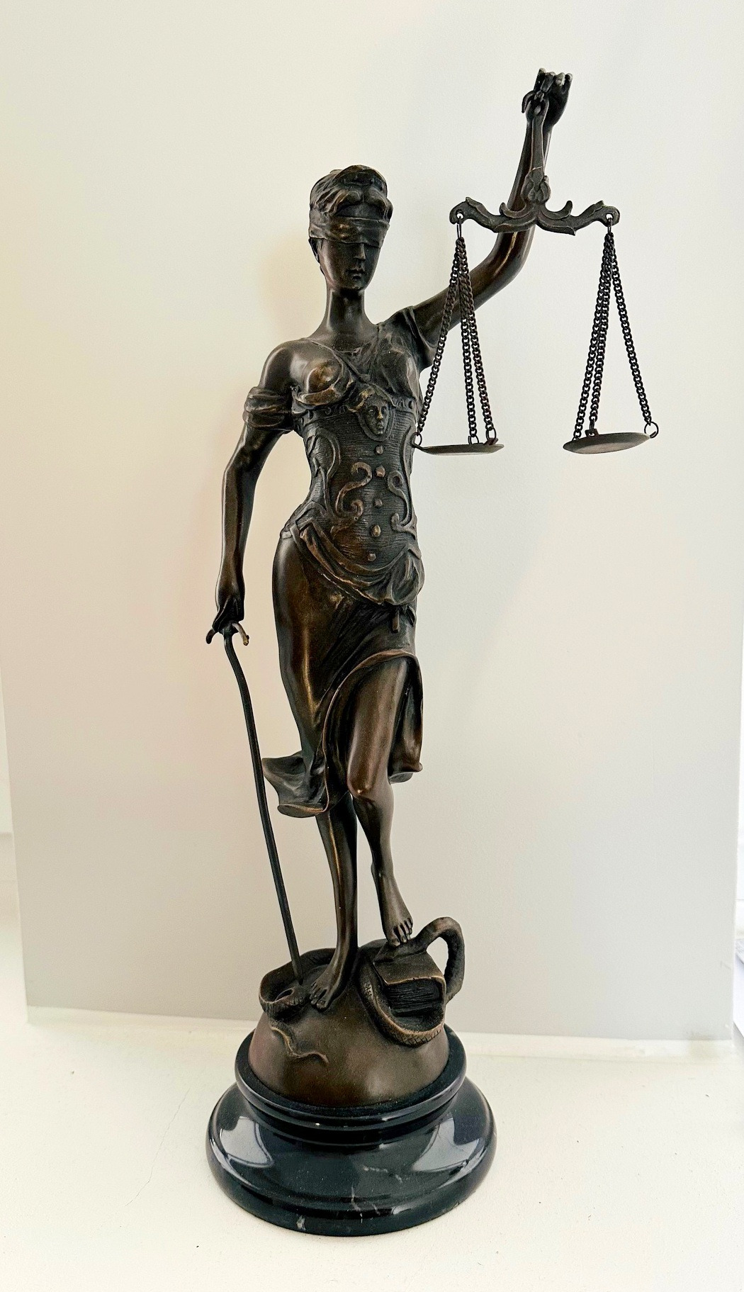 Brązowa statuetka Temidy, bogini sprawiedliwości, z wagą i mieczem, stojąca na postumencie z wężem i książką, na tle białej ściany.