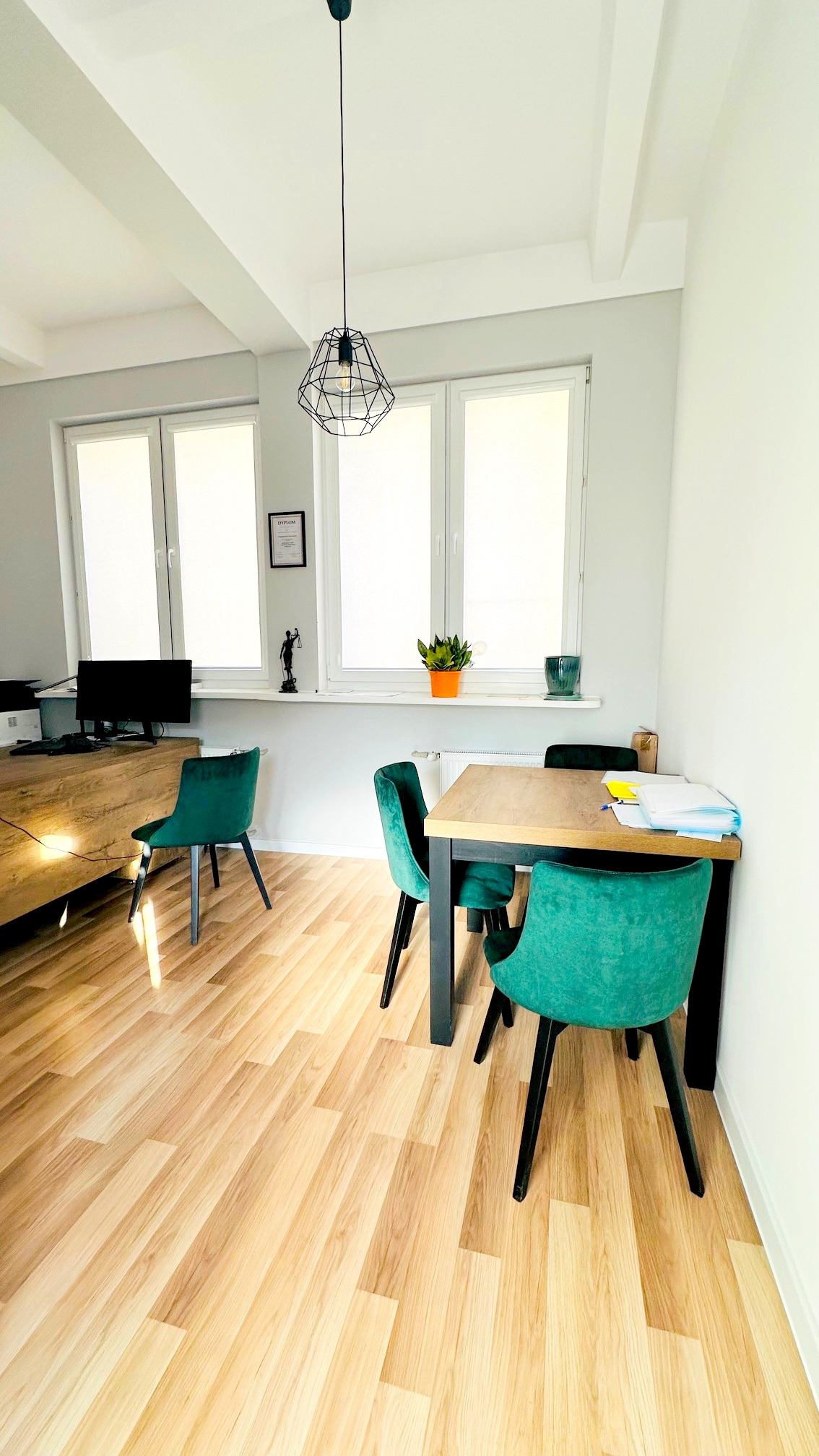 Jasne biuro z drewnianą podłogą, stołem z dokumentami i zielonymi krzesłami, na parapecie figura Temidy i dyplom.