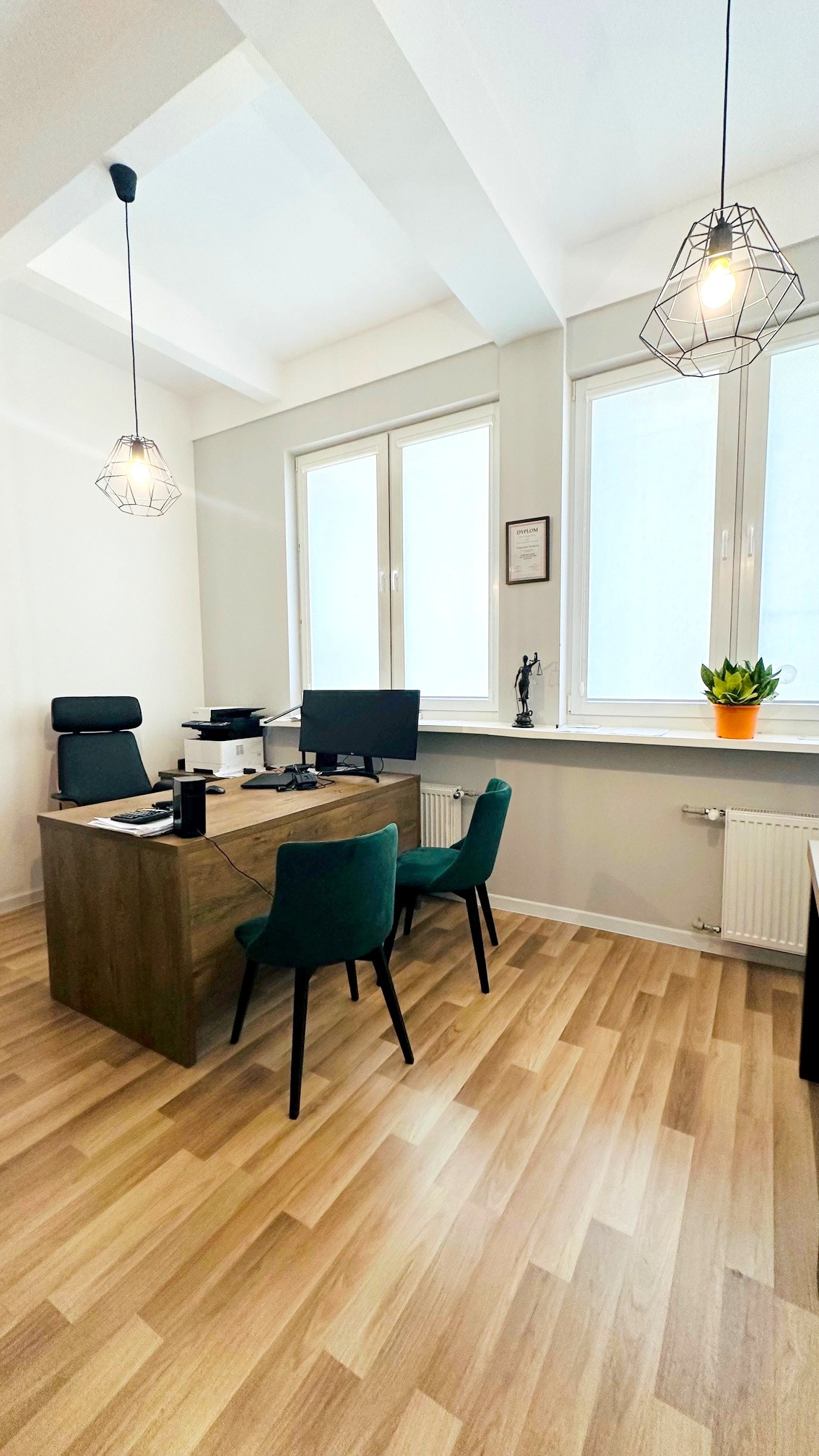 Nowoczesne biuro z drewnianym biurkiem, zielonymi krzesłami i lampami w stylu industrialnym. Na biurku monitor, drukarka i akcesoria biurowe. Na parapecie statuetka Temidy i doniczka z rośliną.