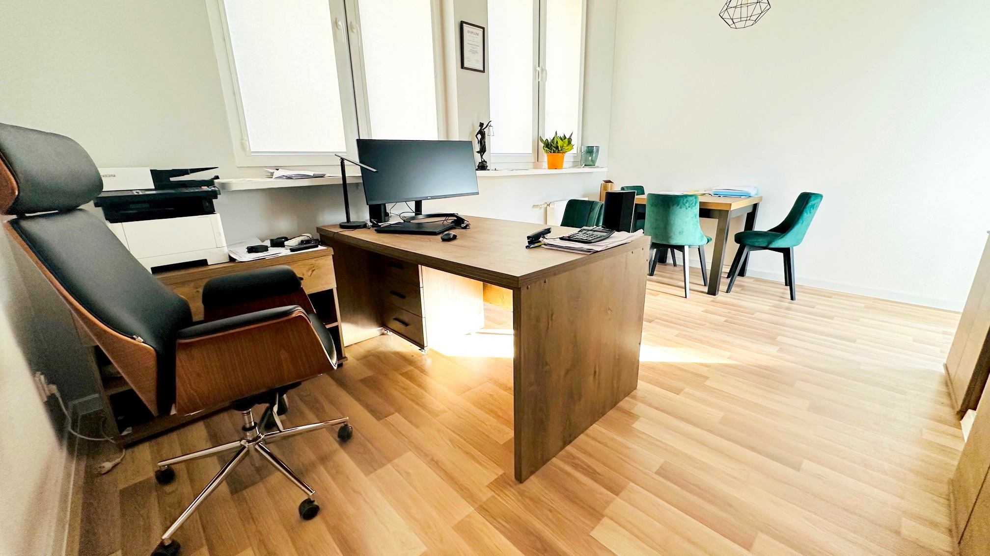 Wnętrze kancelarii w Łodzi: biurko z komputerem, krzesło obrotowe, stół z krzesłami dla klientów, regał z drukarką, na biurku kalkulator i dokumenty.