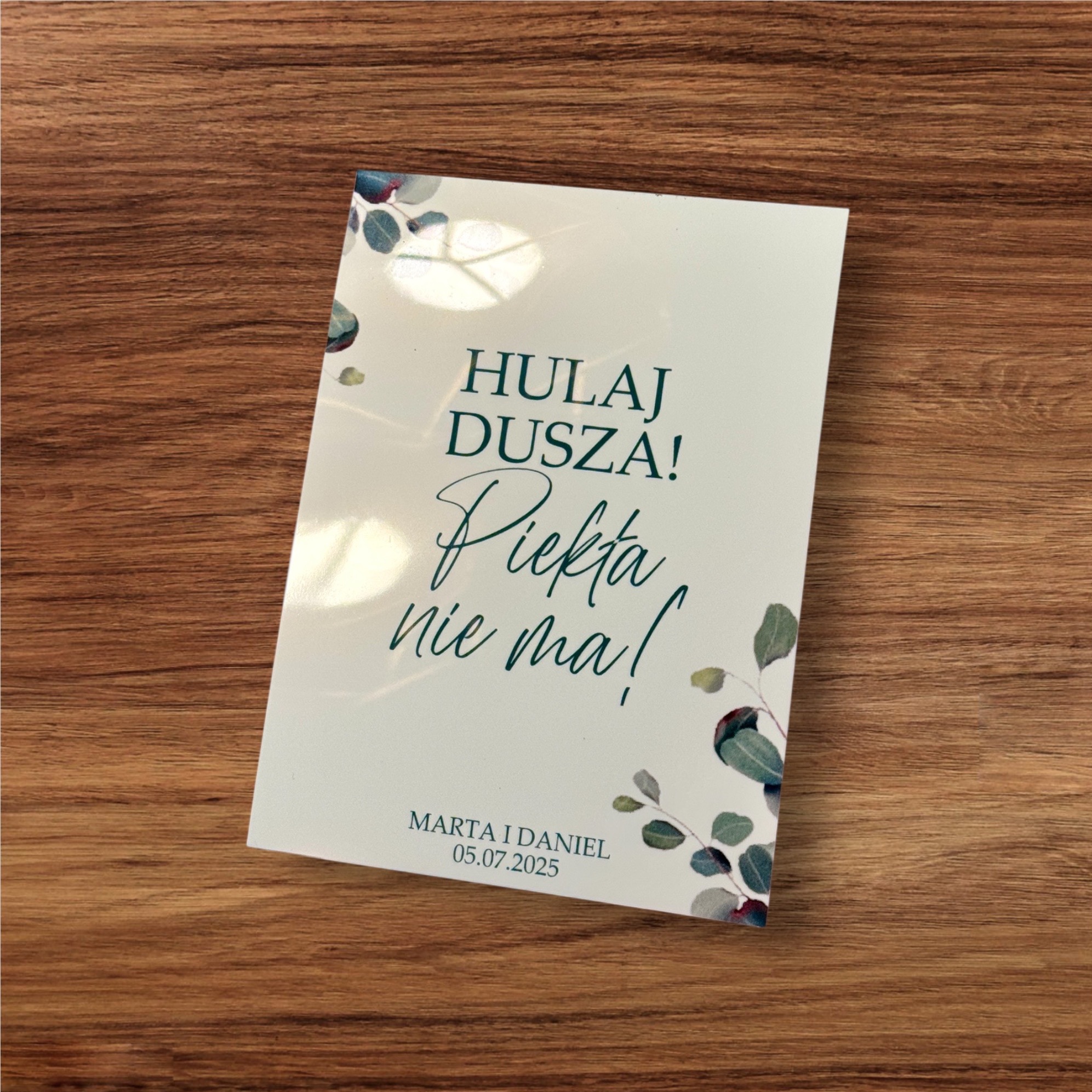 Biała kartka z napisem 'Hulaj Dusza! Piekła nie ma!' i imionami Marta i Daniel oraz datą 05.07.2025, ozdobiona motywem roślinnym, leży na drewnianym stole.
