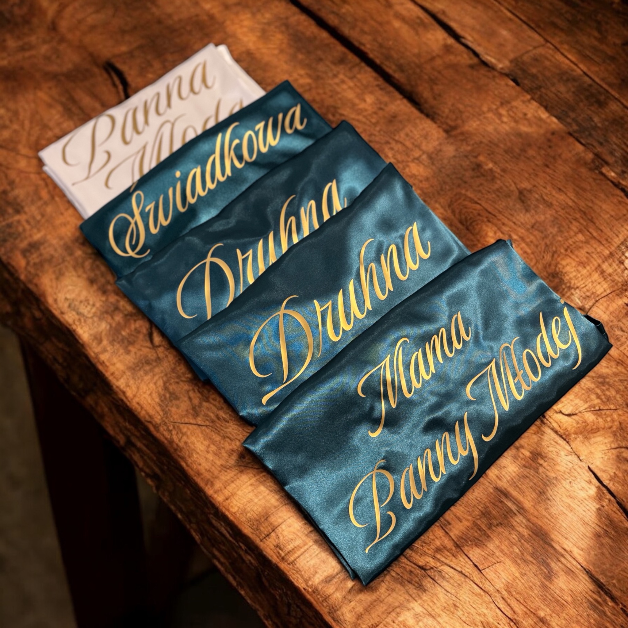 Satynowe szlafroki z imionami i dedykacjami (Panna Młoda, Świadkowa, Druhna, Mama Panny Młodej) w kolorze butelkowej zieleni, leżące na drewnianym blacie.