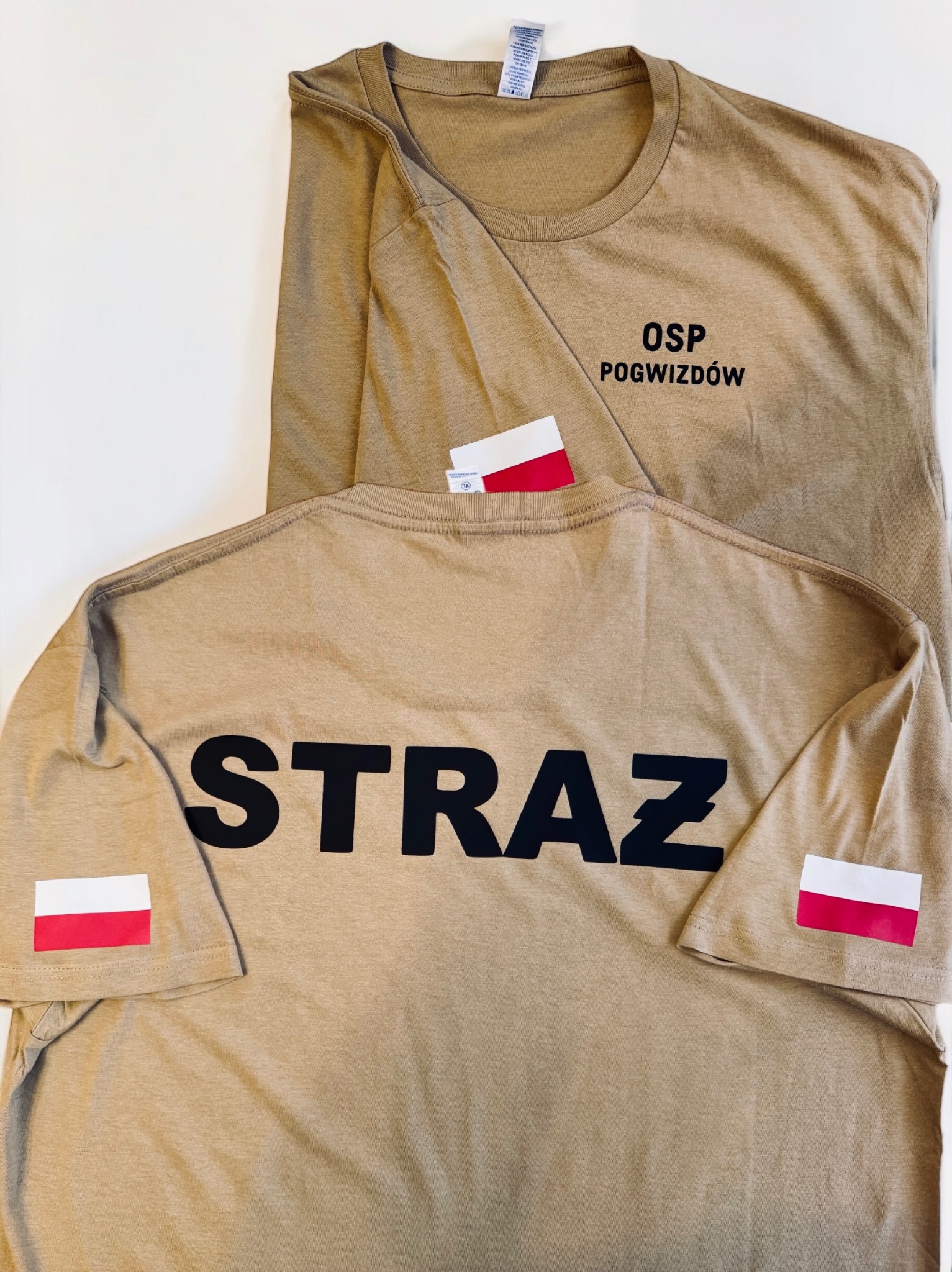 Beżowe koszulki z nadrukiem 'OSP POGWIZDÓW' i 'STRAŻ' oraz flagą Polski na rękawie, leżące na białym tle. Widok z przodu i z tyłu.