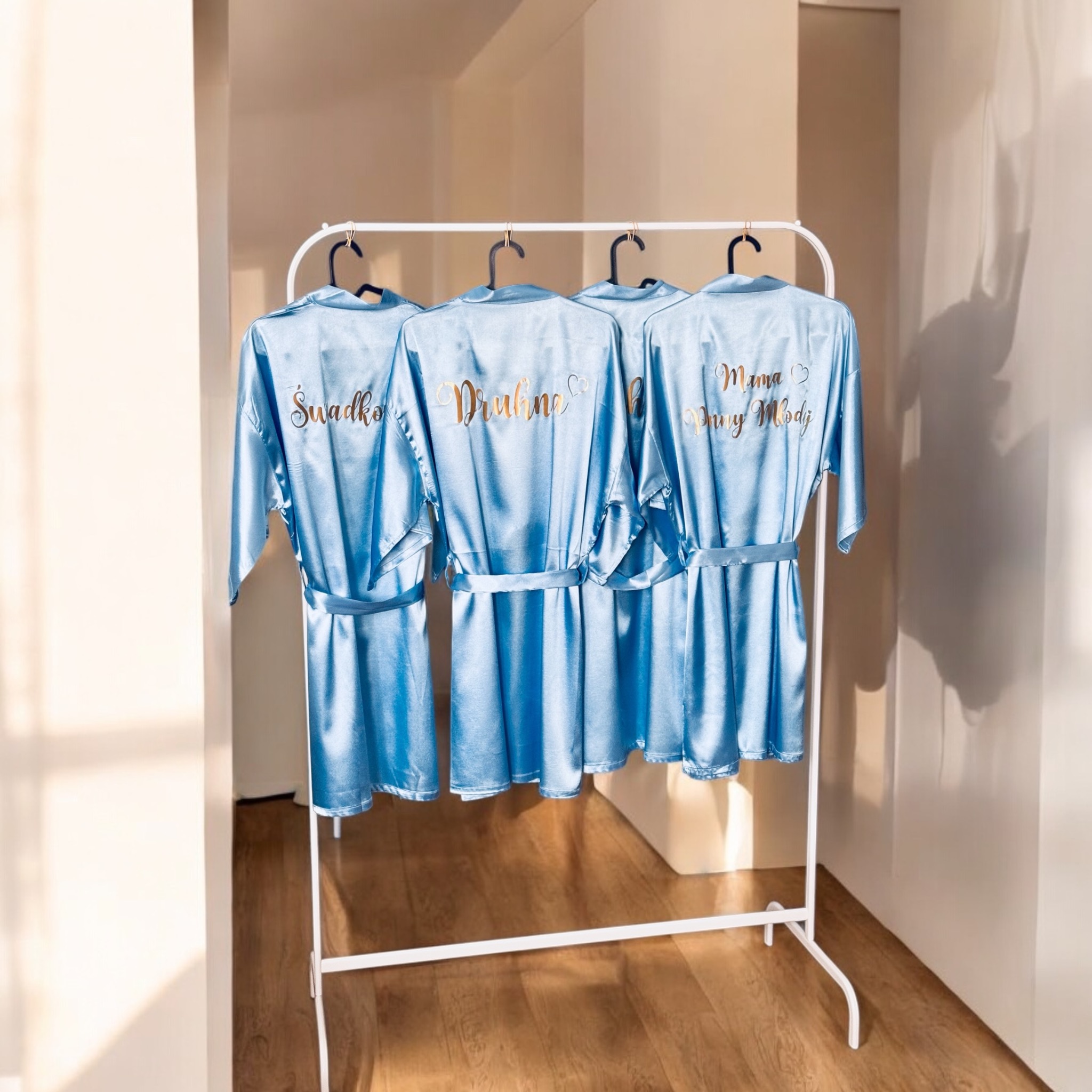 Cztery błękitne, satynowe szlafroki z personalizowanymi, złotymi napisami na wieszaku, ustawione w jasnym, minimalistycznym wnętrzu.