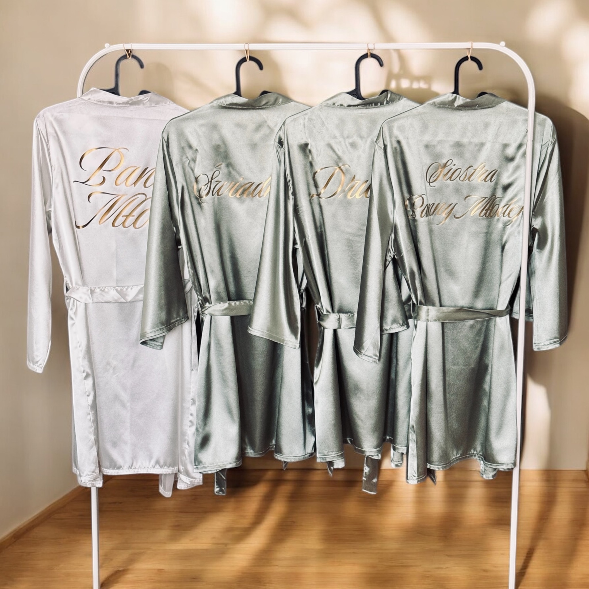Satynowe szlafroki z imionami w kolorze złota, wiszące na wieszaku. Idealne na prezent lub wieczór panieński. Różne kolory i imiona.