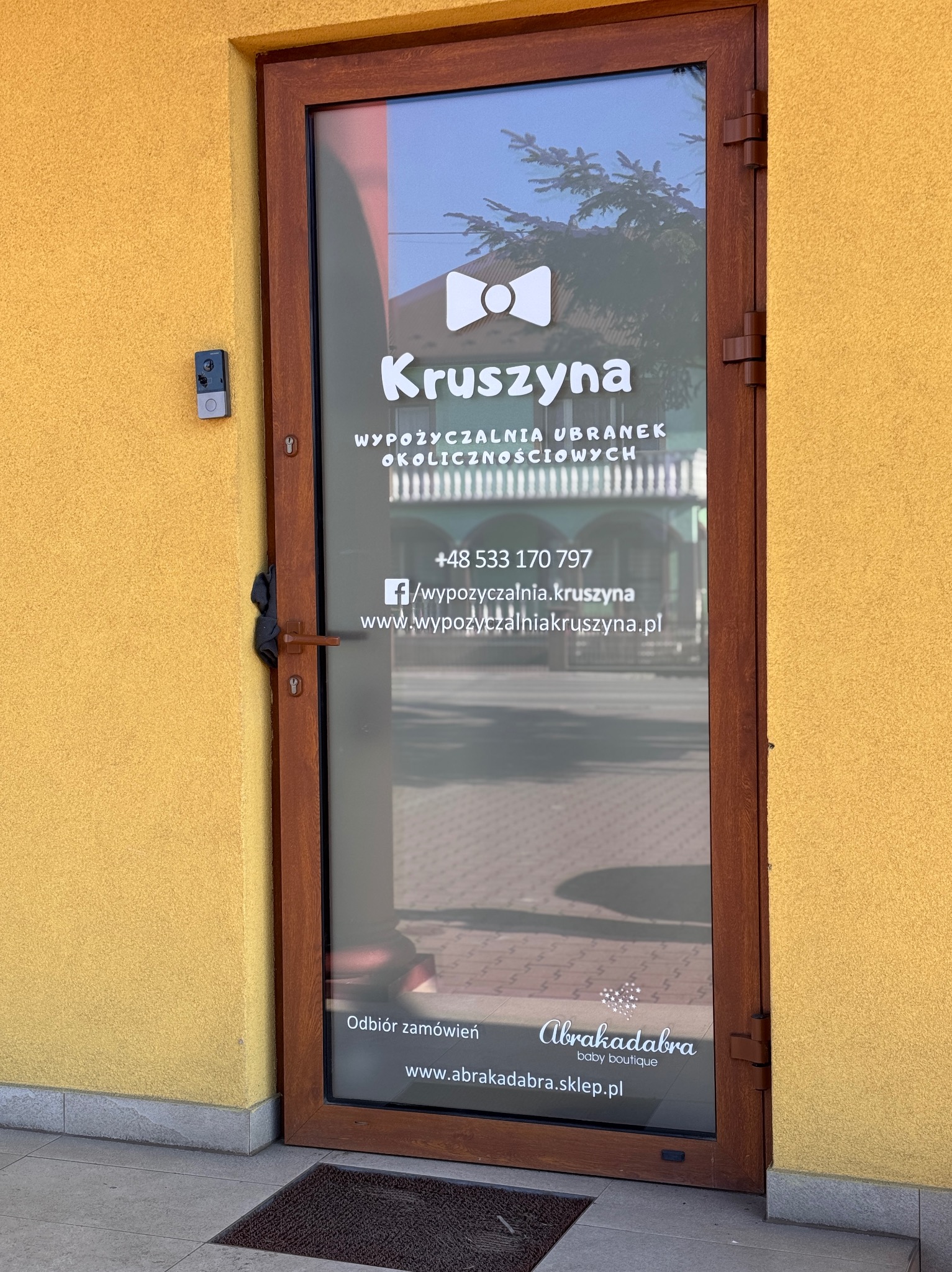 Wejście do wypożyczalni ubrań 'Kruszyna' z naklejonym logo i danymi kontaktowymi na szklanych drzwiach. Widoczny numer telefonu, adres strony www i logo drugiej firmy.