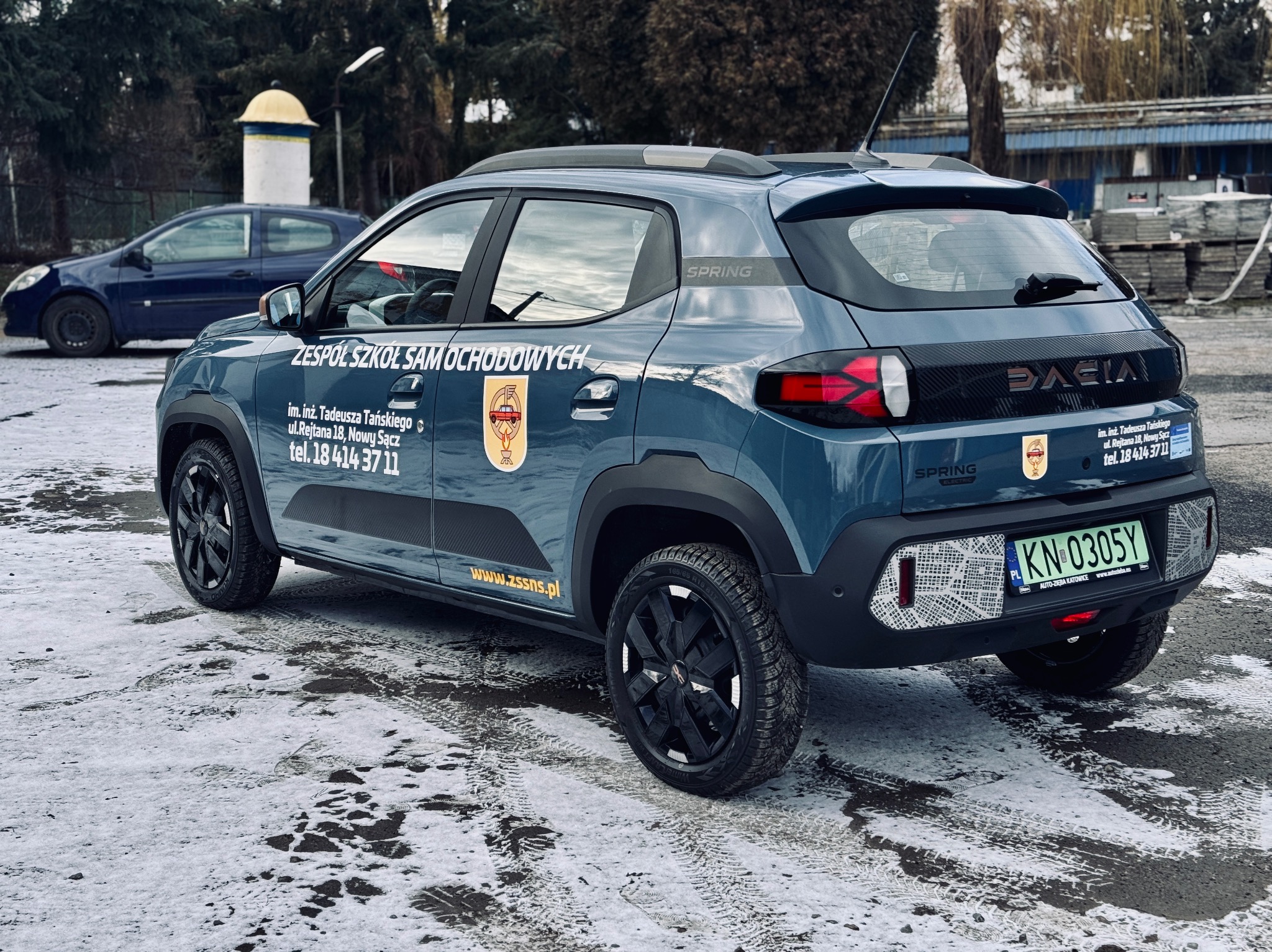 Niebieski samochód Dacia Spring z grafiką szkoły nauki jazdy, widoczny z tyłu i boku na tle zimowego krajobrazu. Na karoserii umieszczone logo i dane kontaktowe szkoły.