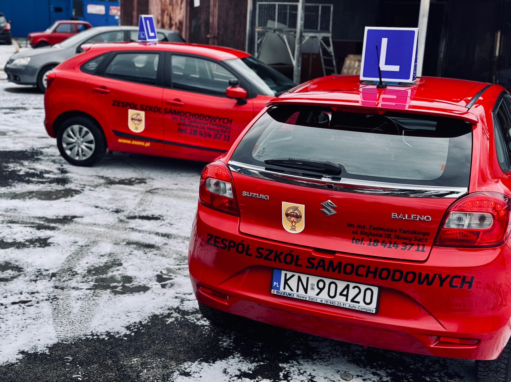 Dwa czerwone samochody Suzuki z oznaczeniem 'L' na dachu, należące do Zespołu Szkół Samochodowych w Nowym Sączu. Widoczne logo i dane kontaktowe na karoserii.