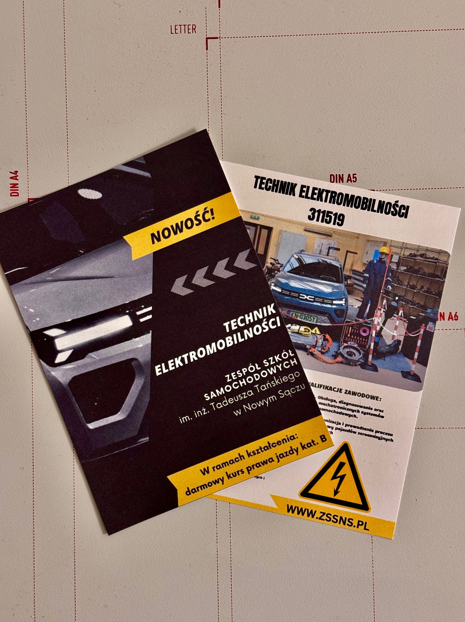 Dwie ulotki reklamowe technikum elektromobilności z informacją o darmowym kursie prawa jazdy kat. B, leżące na brązowym podłożu z podziałką.