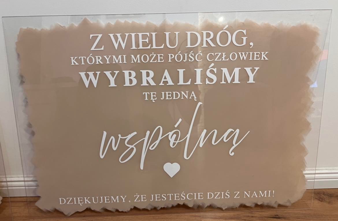 Transparentna tablica z napisem 'Z wielu dróg...' w kolorze białym, umieszczona na beżowym tle. Dekoracja z okazji ślubu lub innego wydarzenia.