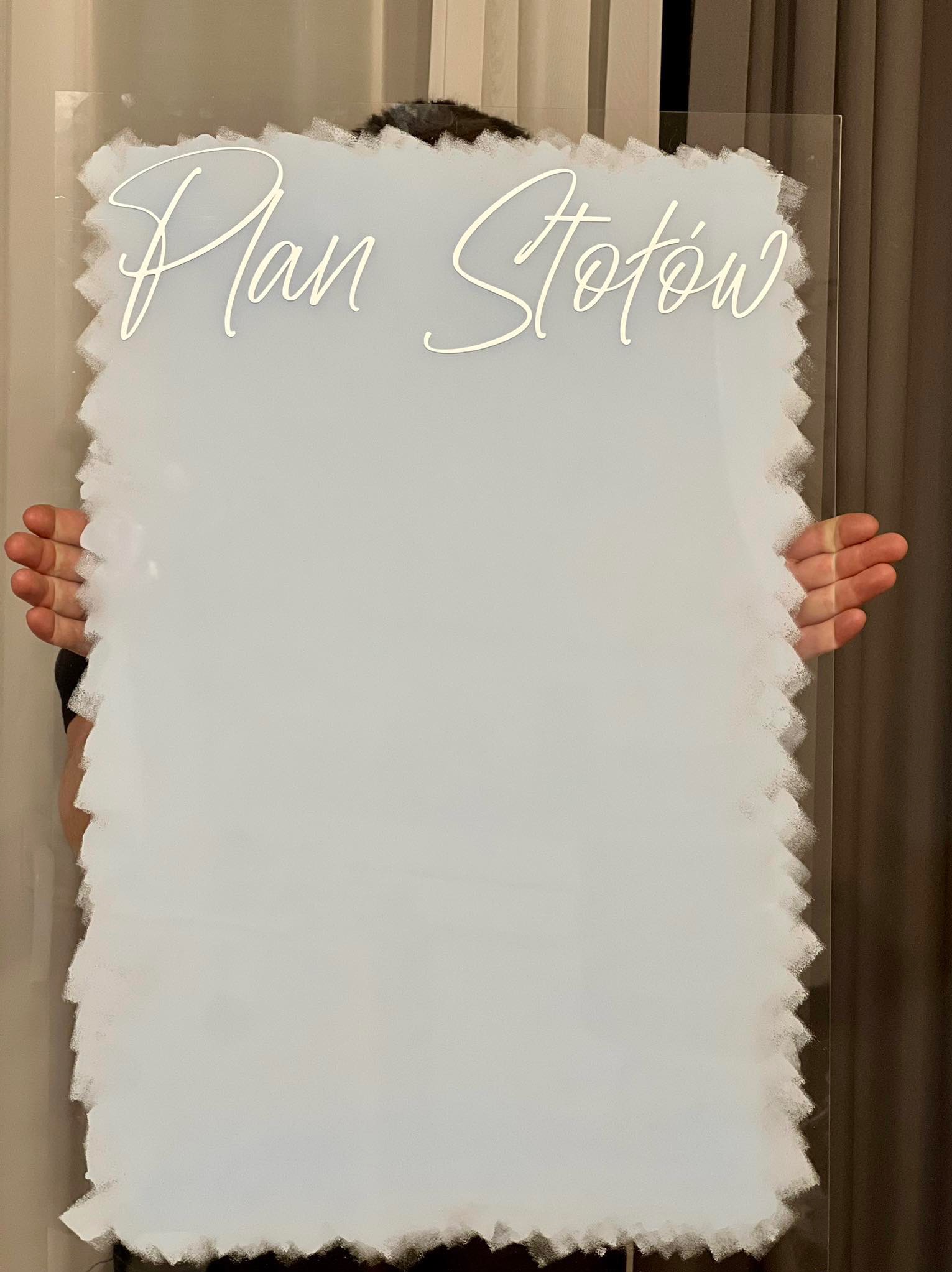 Akrylowa tablica z napisem 'Plan Stołów' w ozdobnej czcionce, trzymana przez osobę. Dekoracja ślubna z unikalnym, nieregularnym obrysem.