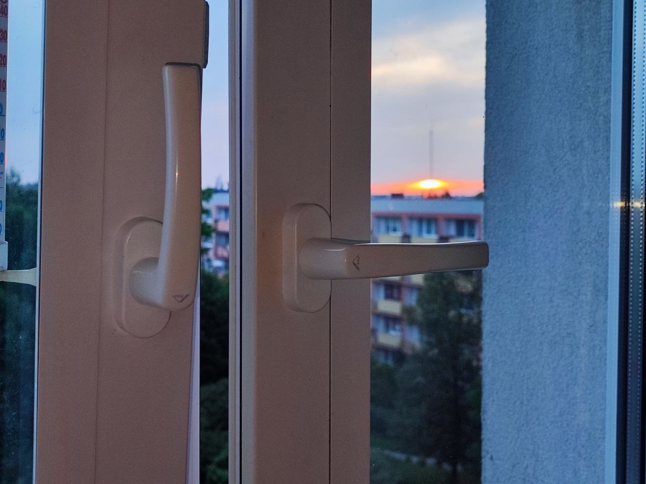 Zamknięte białe okno PCV z widokiem na zachodzące słońce nad blokami mieszkalnymi, widoczny termometr przylepiony do ramy okiennej.