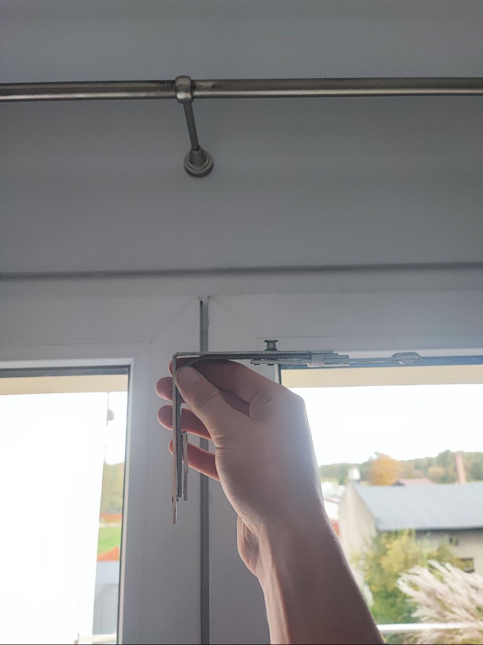 Ręka trzyma metalowy wspornik karnisza okiennego na tle okna i sufitu z zamontowanym karniszem.