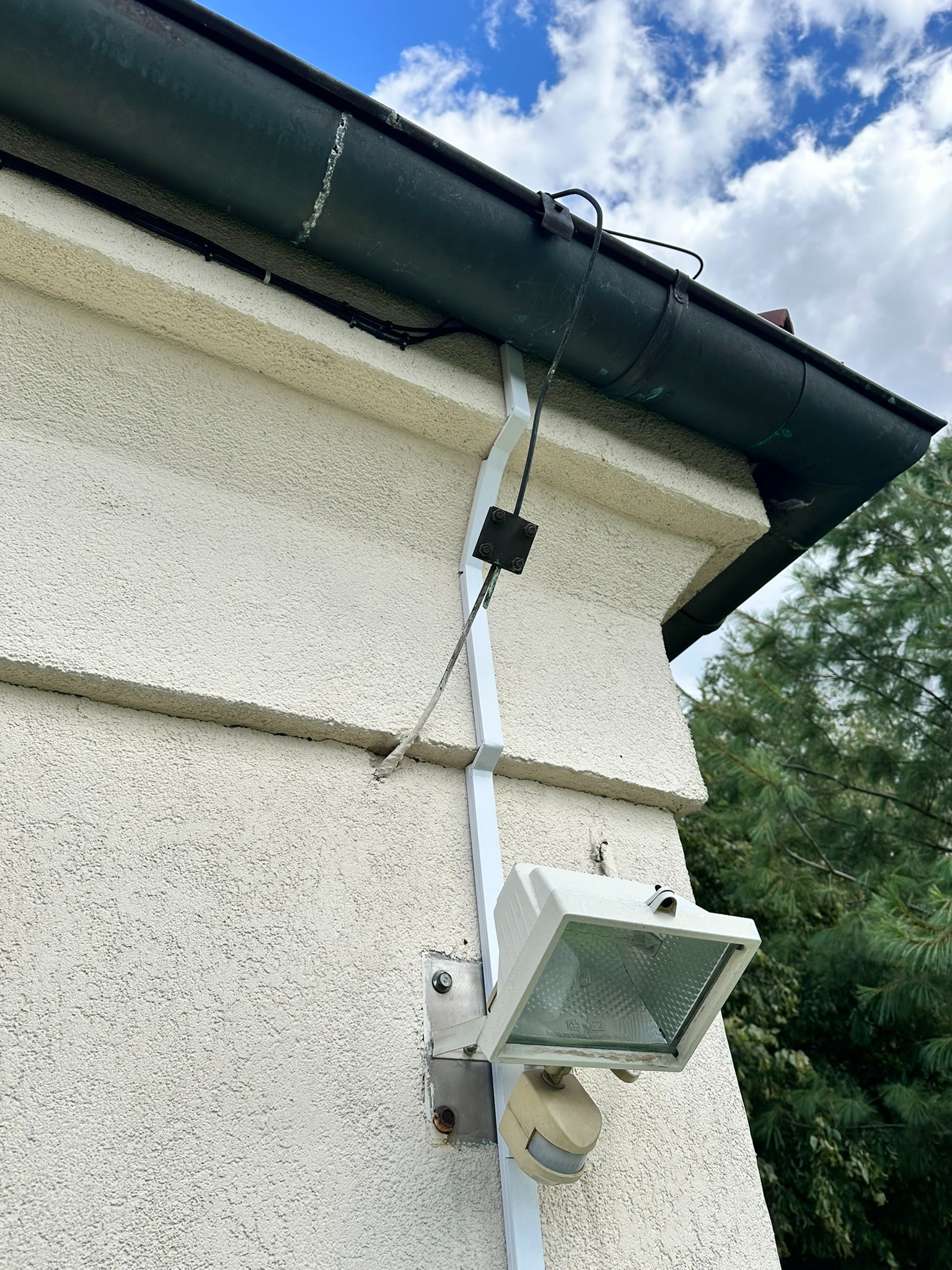 Zewnętrzny reflektor z czujnikiem ruchu, zamontowany na elewacji budynku przy rynnie. Widoczne przewody i elementy mocujące. Biała obudowa lampy na tle jasnej ściany.