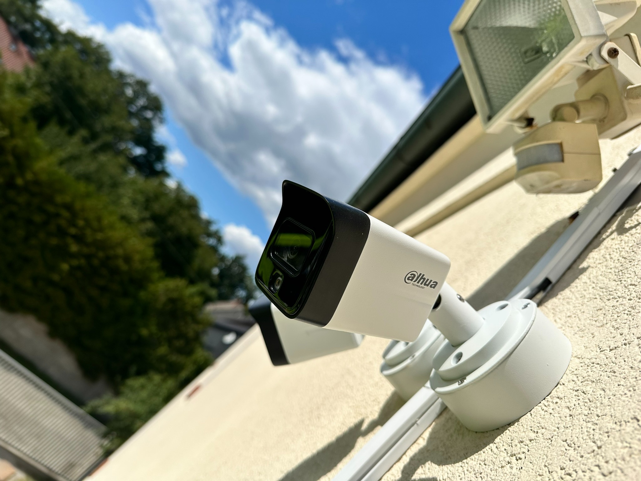 Zewnętrzna kamera monitoringu Dahua na elewacji budynku, z widocznym czujnikiem ruchu i fragmentem nieba z chmurami w tle. Montaż systemu alarmowego.