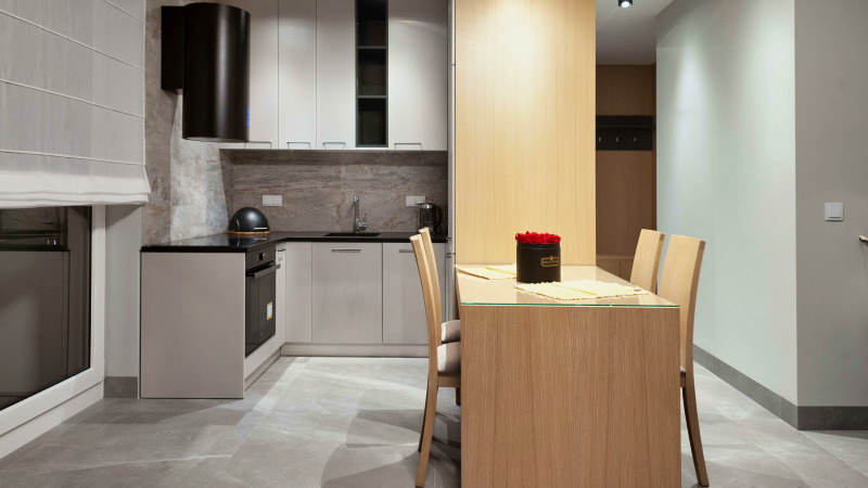 Jasna, nowoczesna kuchnia z szarymi płytkami, białymi szafkami i czarnym okapem. Na pierwszym planie stół z drewnianym blatem i krzesłami. Na stole czarny pojemnik z czerwonymi różami.