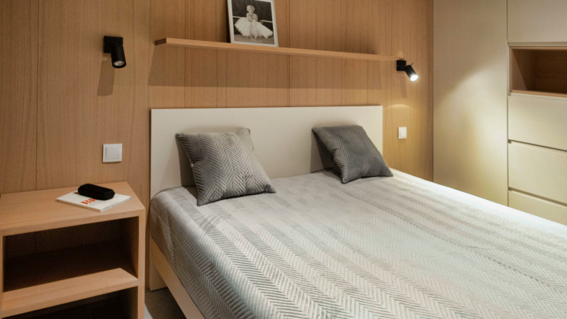 Jasna sypialnia z drewnianą boazerią, łóżkiem z szarą pościelą w jodełkę i szafką nocną z lampką oraz półką z czarno-białym zdjęciem.