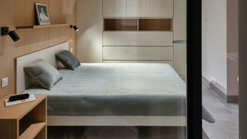 Nowoczesna sypialnia z łóżkiem, szafką nocną i dużą szafą w zabudowie z połyskującymi frontami.