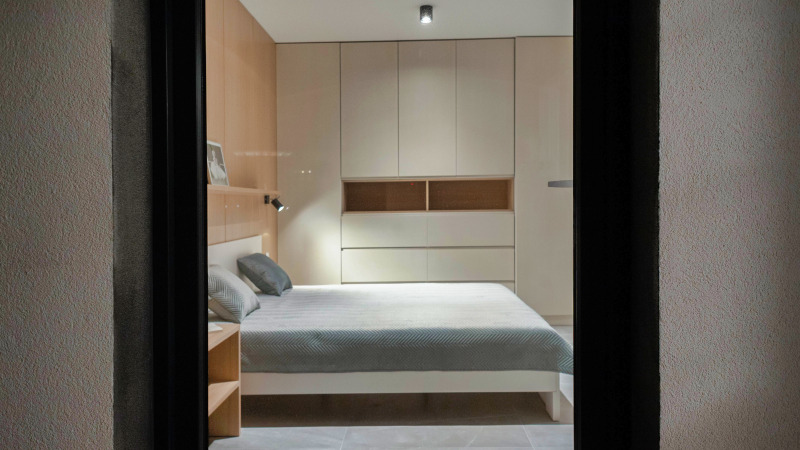 Nowoczesna sypialnia z łóżkiem i zabudową meblową w odcieniach beżu, widok przez framugę drzwi.