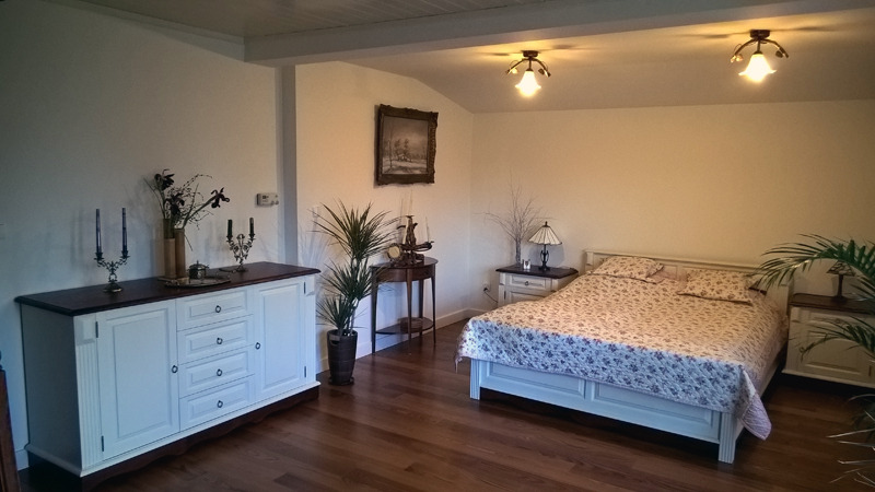 Jasna sypialnia z drewnianą podłogą, białą komodą z ciemnym blatem, łóżkiem z kwiecistą pościelą i roślinami doniczkowymi.