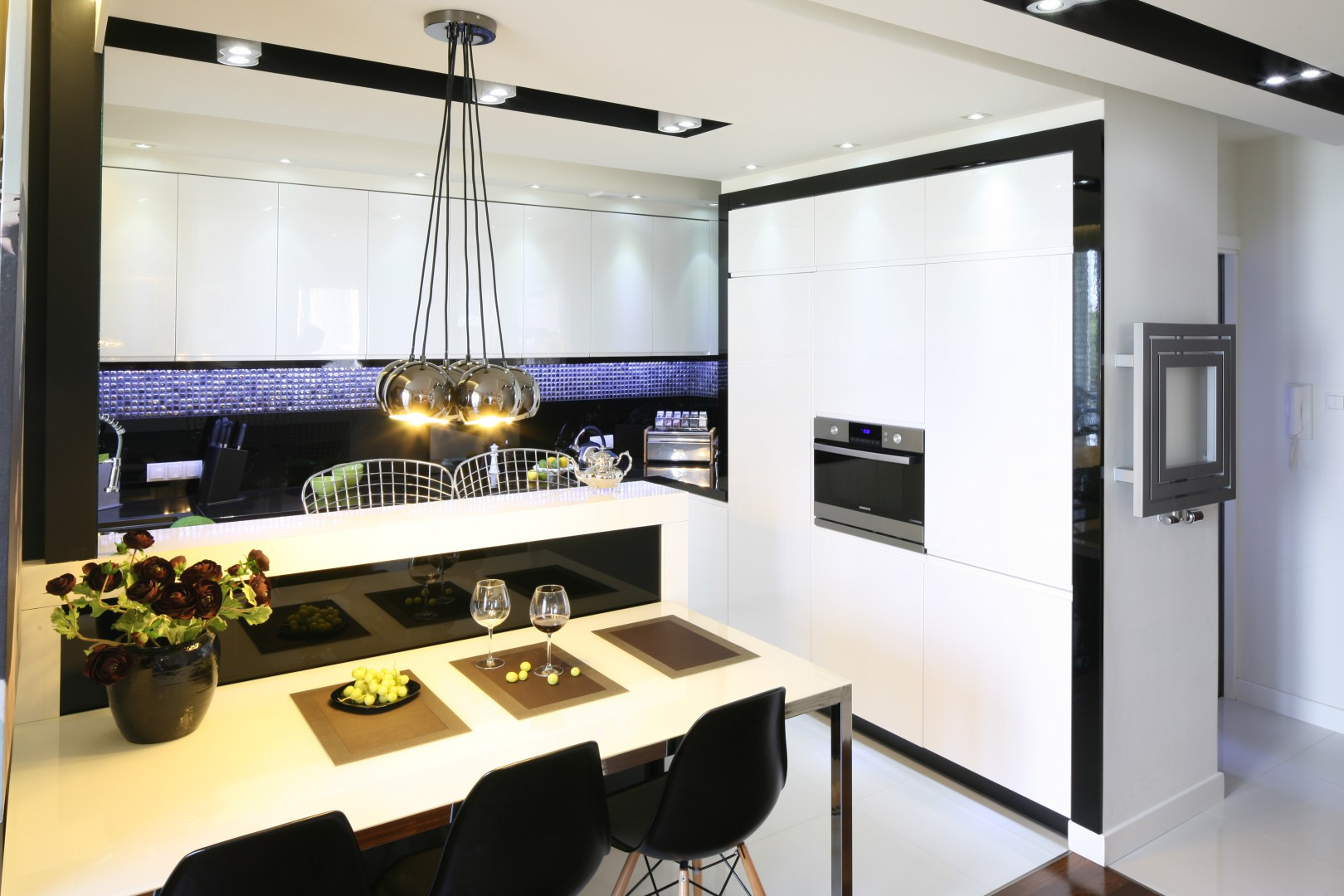 Nowoczesna kuchnia z białymi, błyszczącymi frontami, czarnymi akcentami i szklanym panelem nad blatem, oświetlona wiszącą lampą z kilkoma kloszami, z widocznym stołem z owocami i kieliszkami.