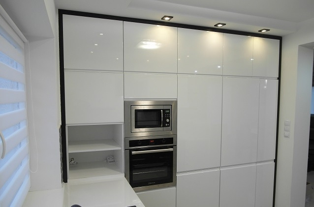 Biała, lakierowana kuchnia na wymiar z czarnymi obramowaniami, zabudowana z piekarnikiem i mikrofalówką, oświetlenie punktowe w suficie, roleta okienna.